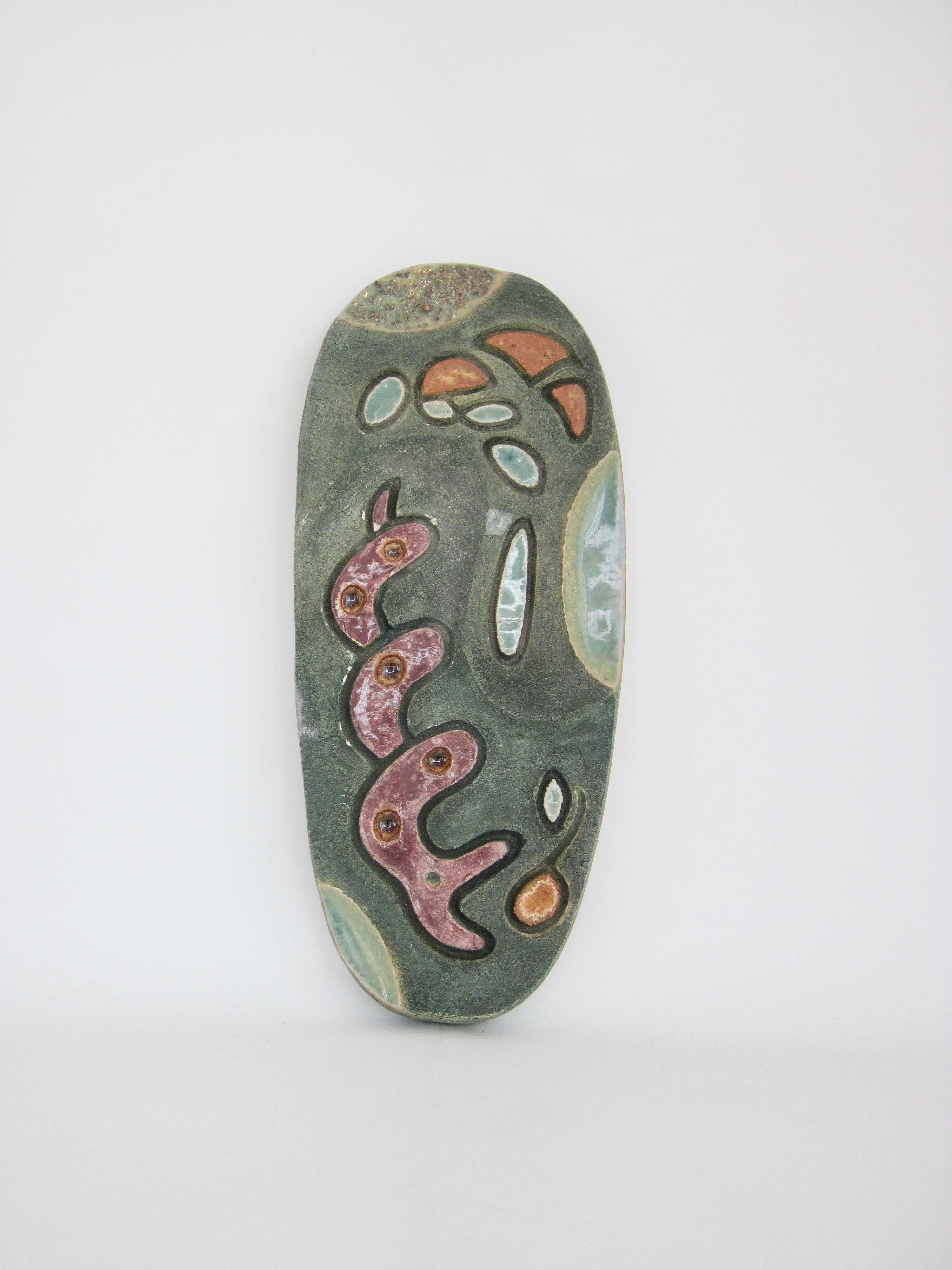 Studio made artwork drawn from organic shapes into decorative form, this spectacular green oval ceramic slab sculpture with dark hued incised outlines around colorful abstract shapes evokes a natural environment, movement, and it's continual change. 