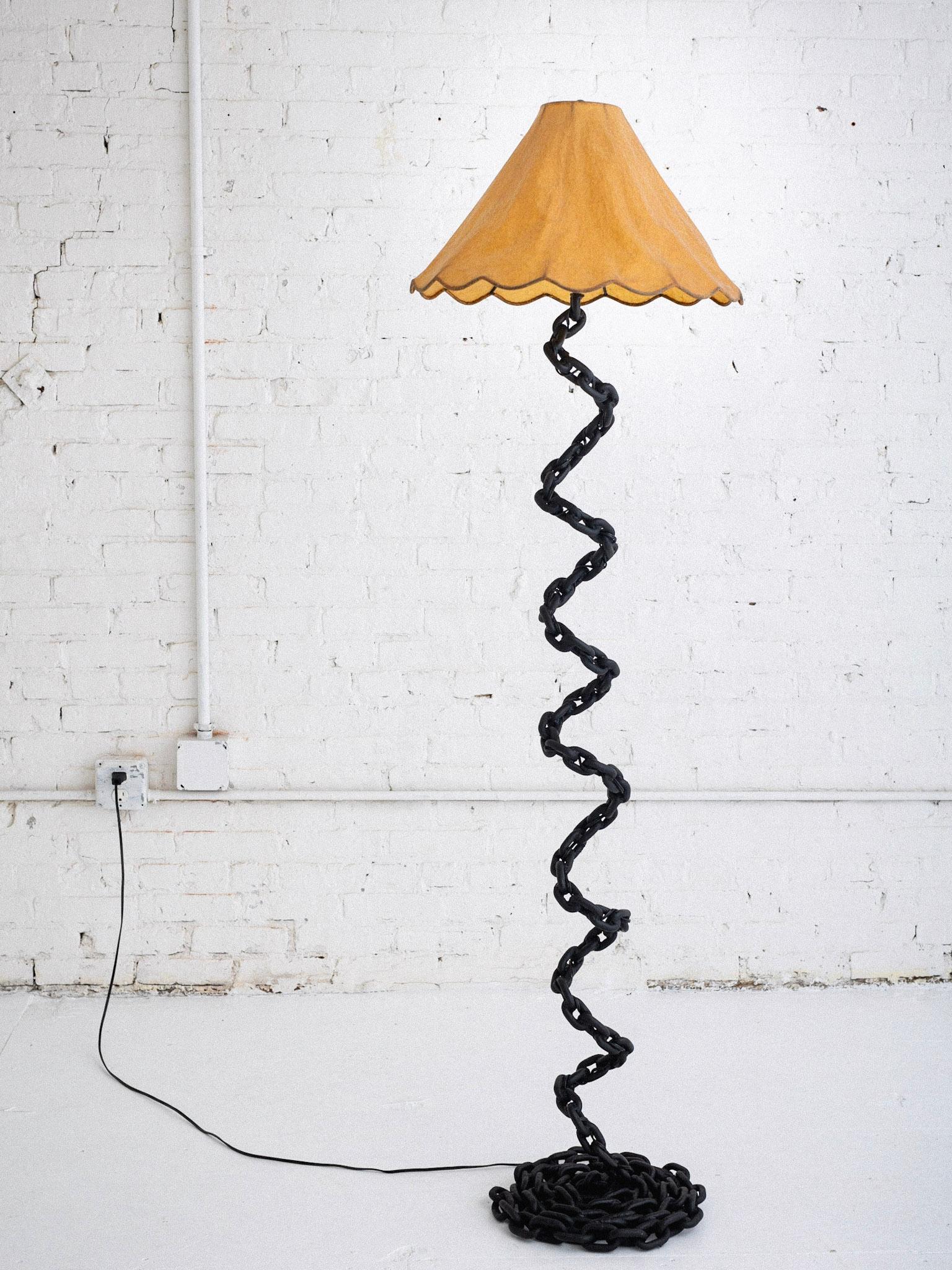 A studio made chain link floor lamp. Hand welded links spiral to create a twisted silhouette on a coiled chain base. Painted black. One available. Shade not included. Measurements are from the base to the top of the socket. The shade harp adds an