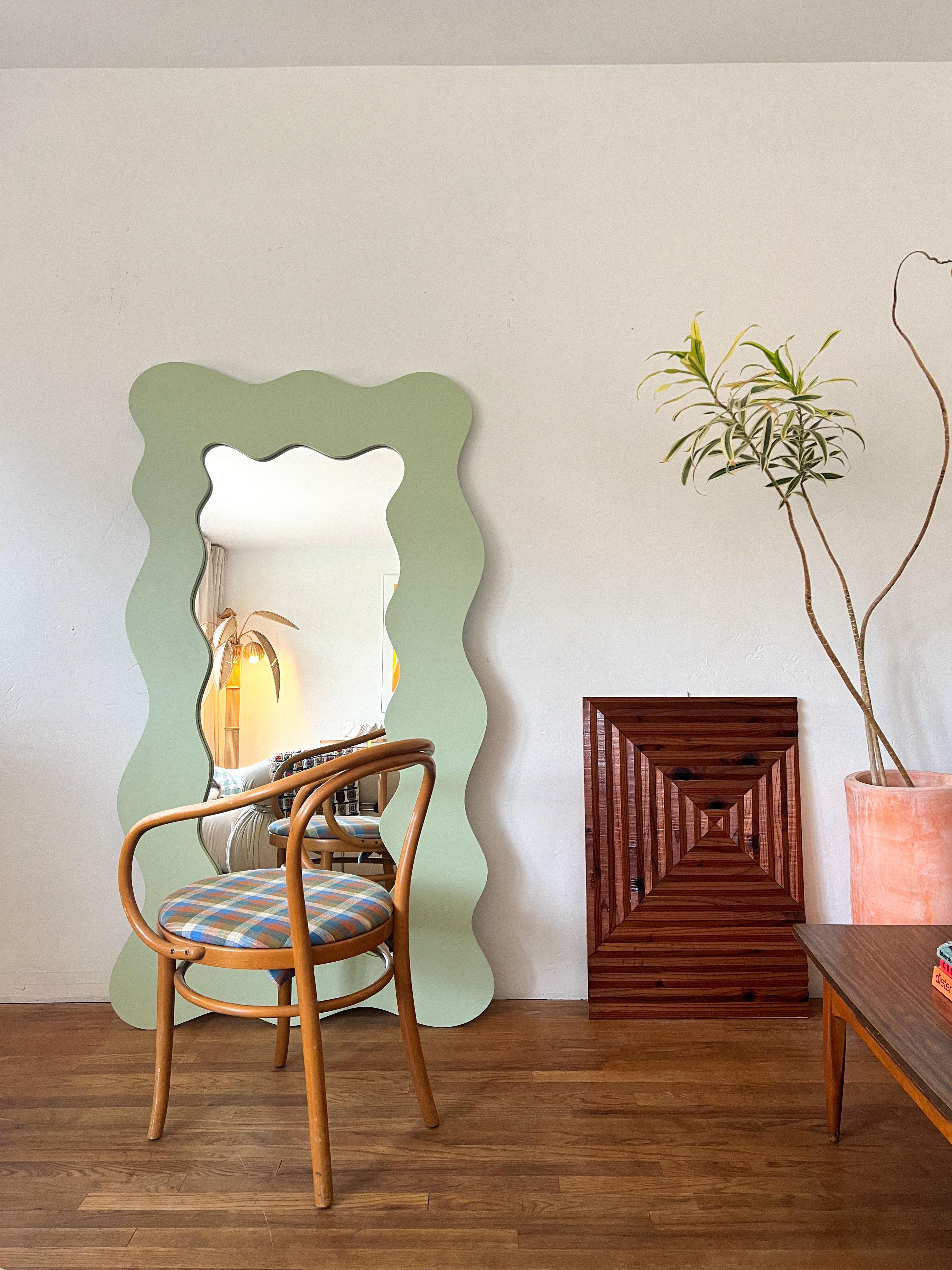 Un miroir de sol audacieux et sculptural doté d'un cadre ondulant à bord ondulé dans une douce finition vert sauge. Cette pièce saisissante s'inspire du mouvement emblématique Memphis Milano des années 1980 et du renouveau du design postmoderne