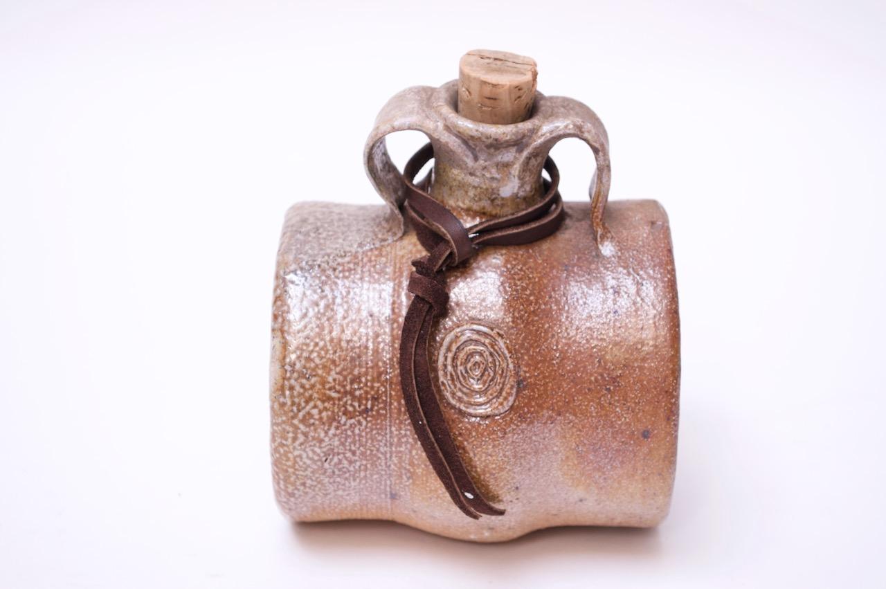 1973 Pollack studio stoneware whiskey nip / barrel with cork. Attractive partially metallic brown / taupe color combination with mottled surface. Includes a decorative leather tie and original cork. 
Incised 