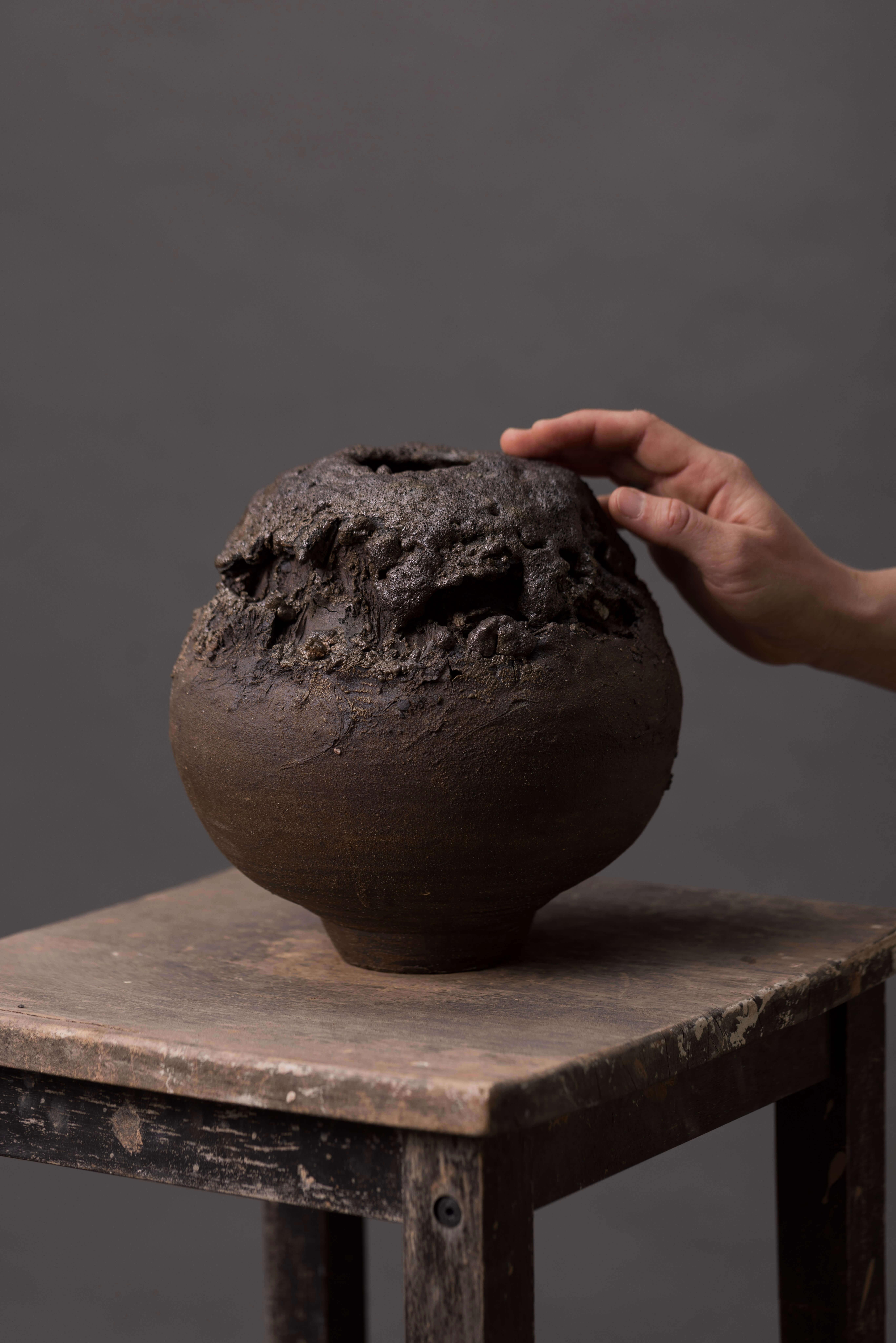 Questo vaso in ceramica lavorato a mano ha una forma rotonda e una superficie marrone scuro caratterizzata da una texture irregolare e ruvida. L'esterno presenta buche, fessure e piccoli fori che creano un aspetto tattile ispirato alla roccia