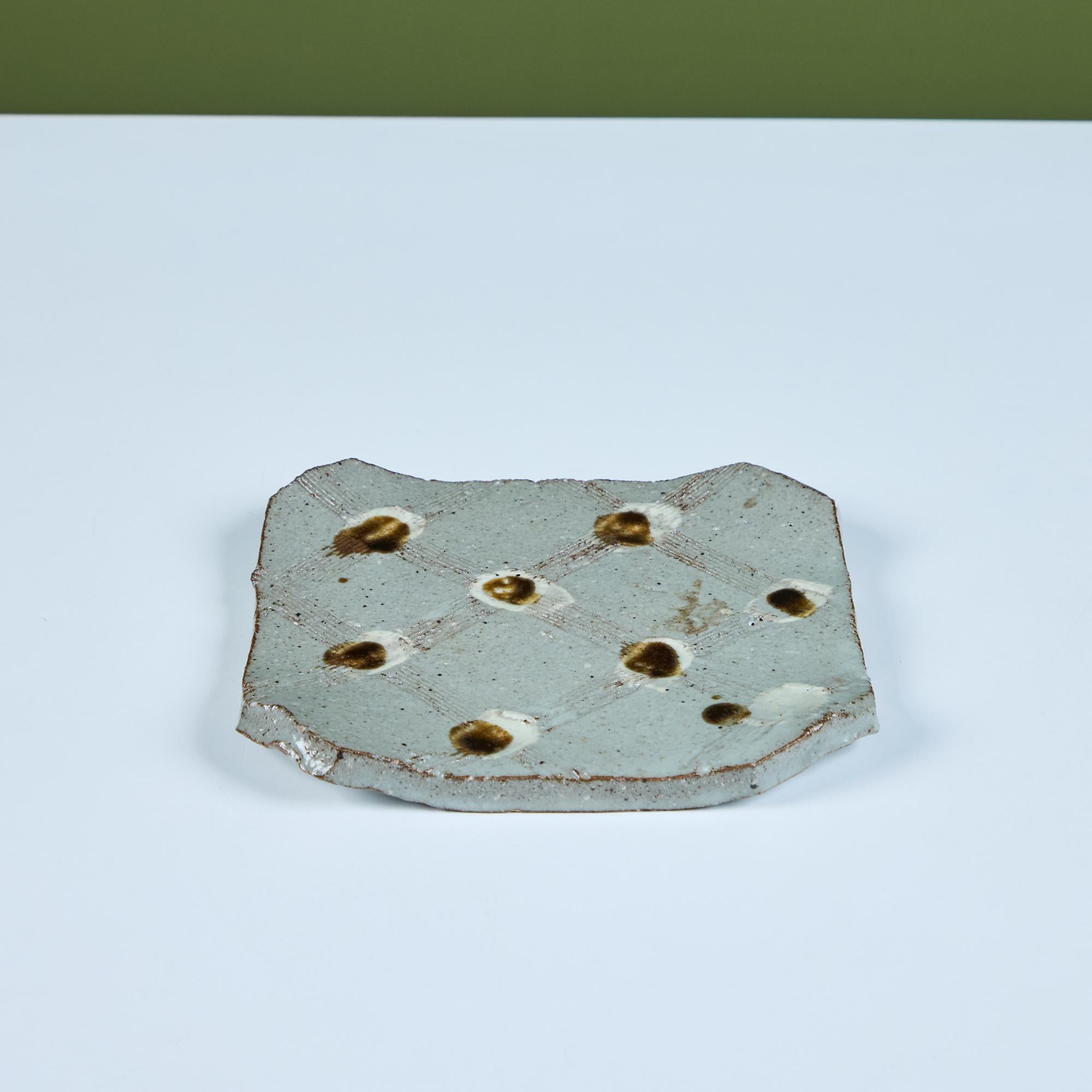 Plato de cerámica con esquinas torneadas hacia arriba y esquinas escuadradas. El plato hexagonal presenta ocho surcos esmaltados de color marrón poco profundos en la superficie del plato, con líneas incisas que conectan los puntos formando un dibujo