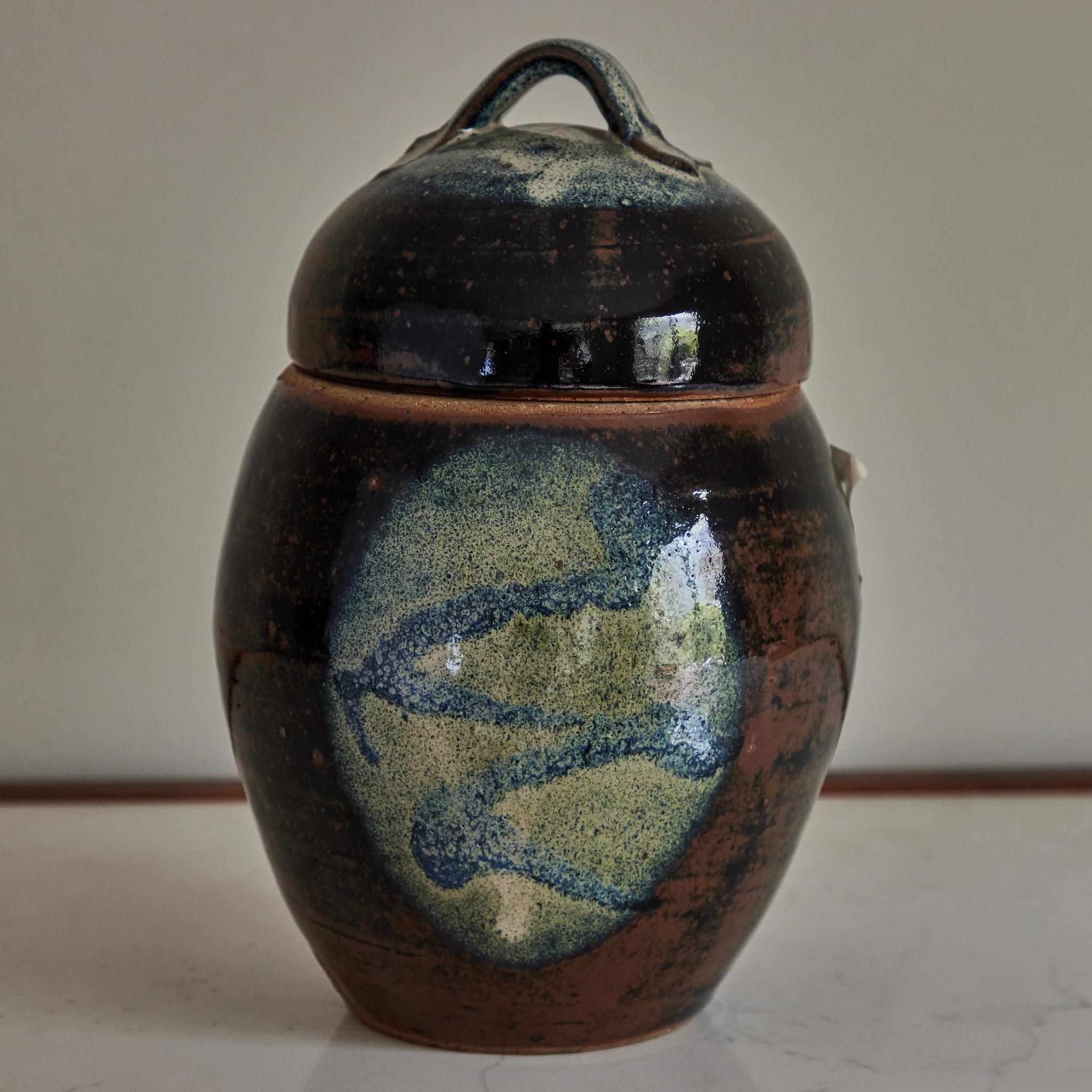 English mid-century studio pottery jar with handle-topped lid. A fabulous mix of pale mossy greens and aquatic blues, with washes of rich loamy browns and volcanic black, the jar has an earthy, elemental quality. 

England, circa 1960

Dimensions:
