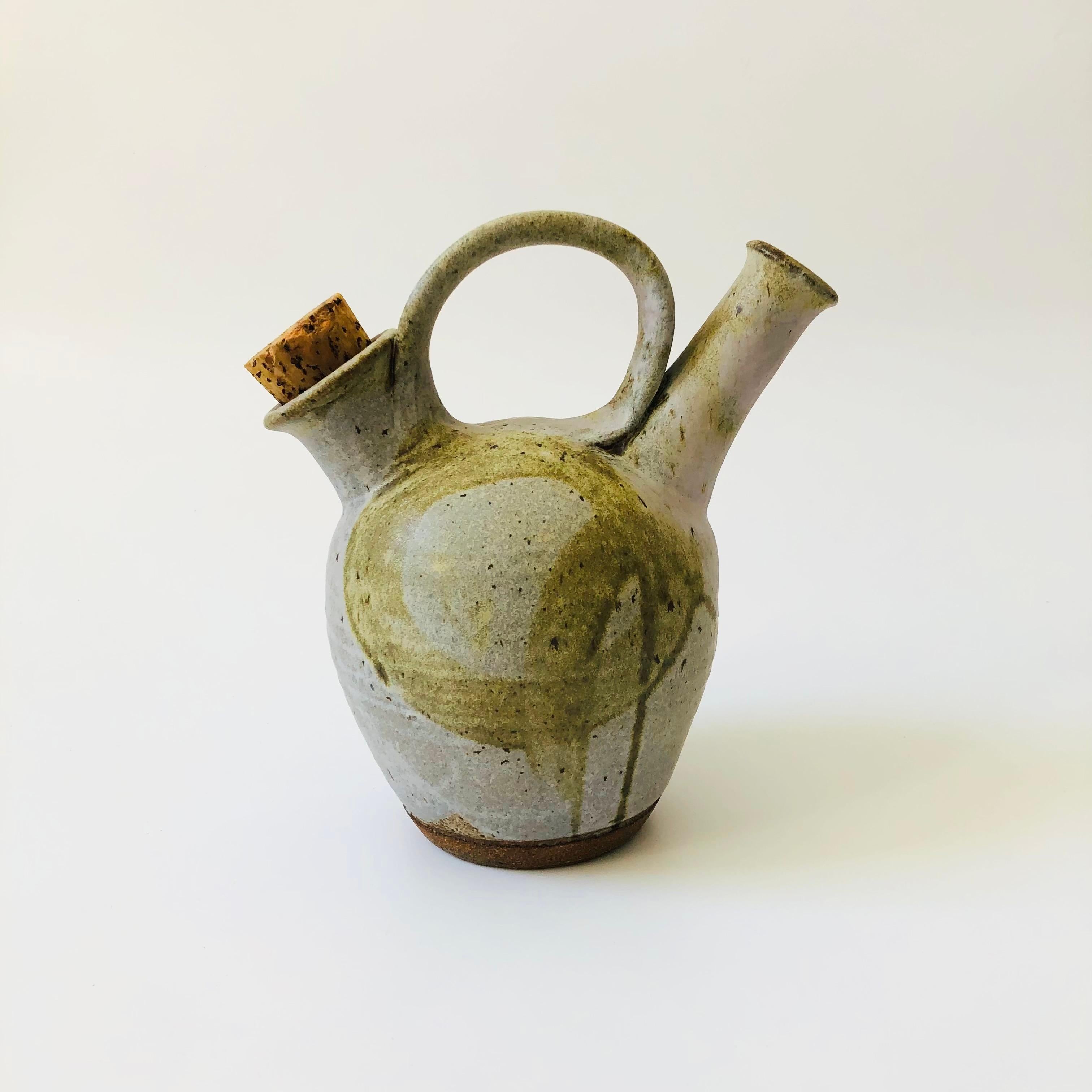 A vintage studio pottery pitcher. Lovely speckled gray glaze with a freeform hand painted design in the center. Features a center handle with a spout on one side and a corked opening for refilling on the other side. Maker is unknown, signed 
