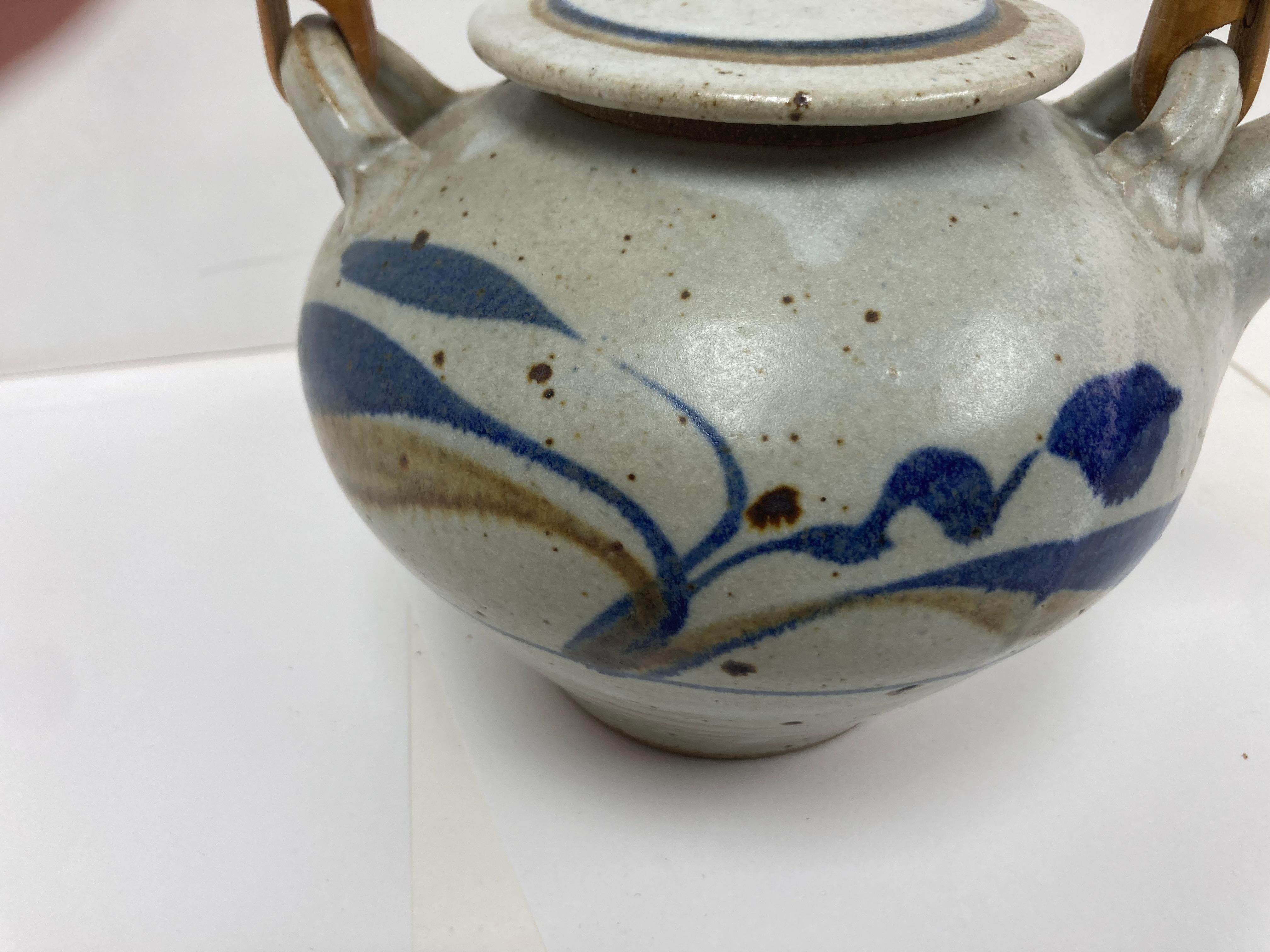 A fine studio pottery Tea Server with fine form and craftsmanship. The pot with decorative free form foliate decoration in brown and blue on a pleasing spotted oyster background and having  a fine soft polished glaze. The handle is bent rattan with
