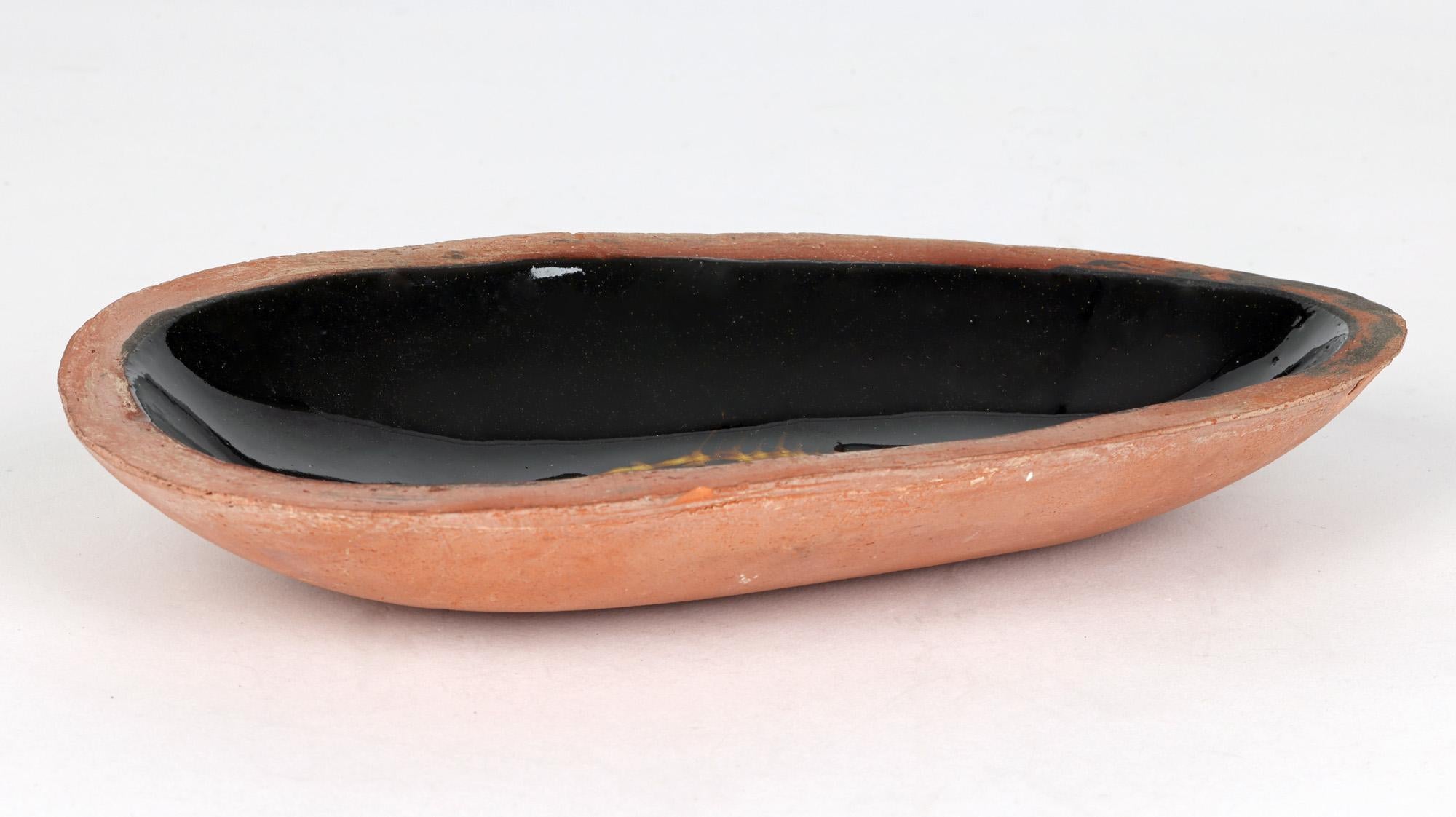 A stylish British studio pottery dish decorated with a slipware fish design in the manner of Winchcombe and signed Gilbert dating from the 20th century. The oval shaped dish narrows to one end and is heavily hand made from thick terracotta clay with