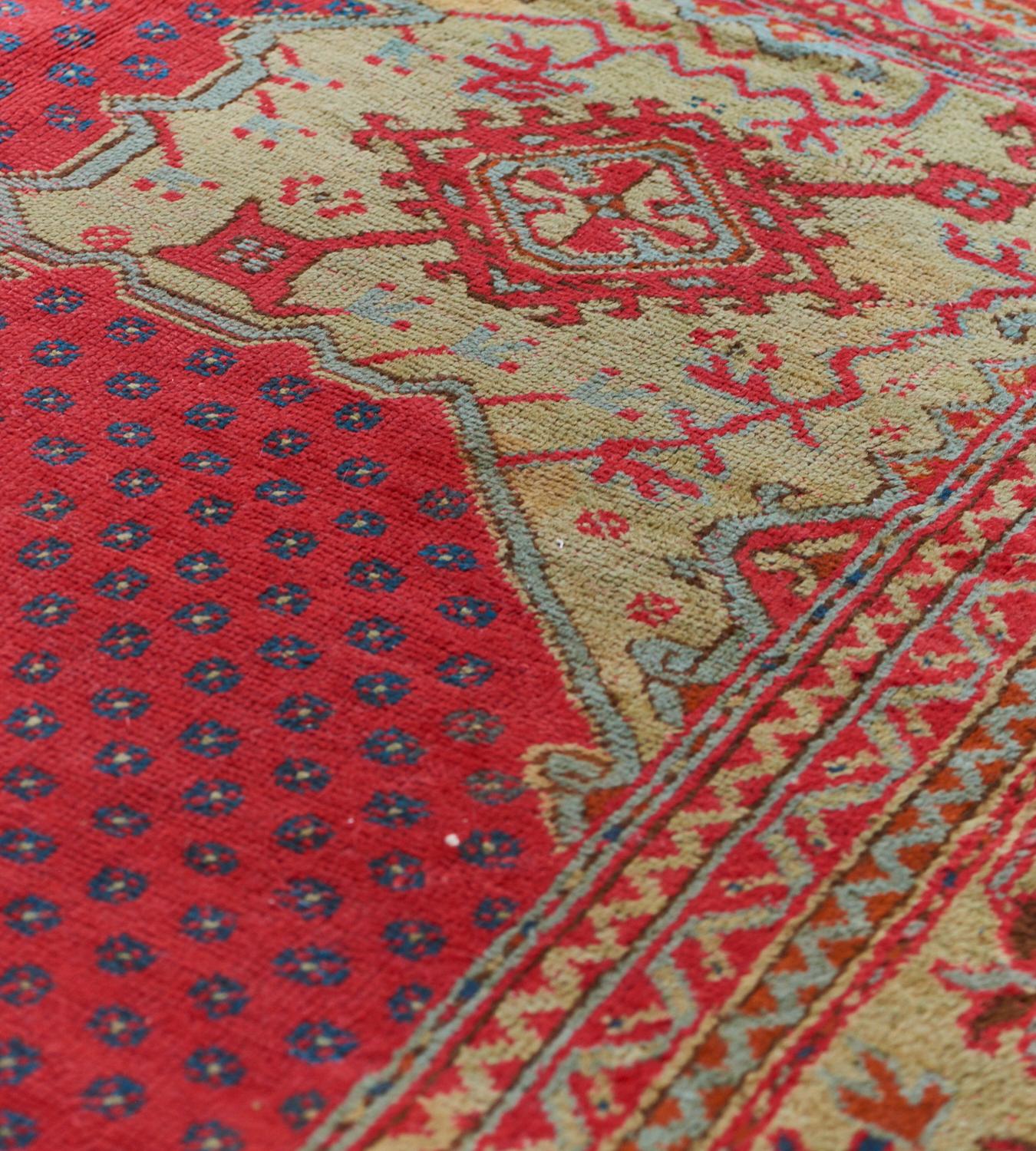 This antique Oushak rug has a tomato-red field scattered with tiny blue roundels around a central pistachio-green and light blue lozenge medallion with a cherry-red central lozenge issuing angular floral vine and shield lozenges and further