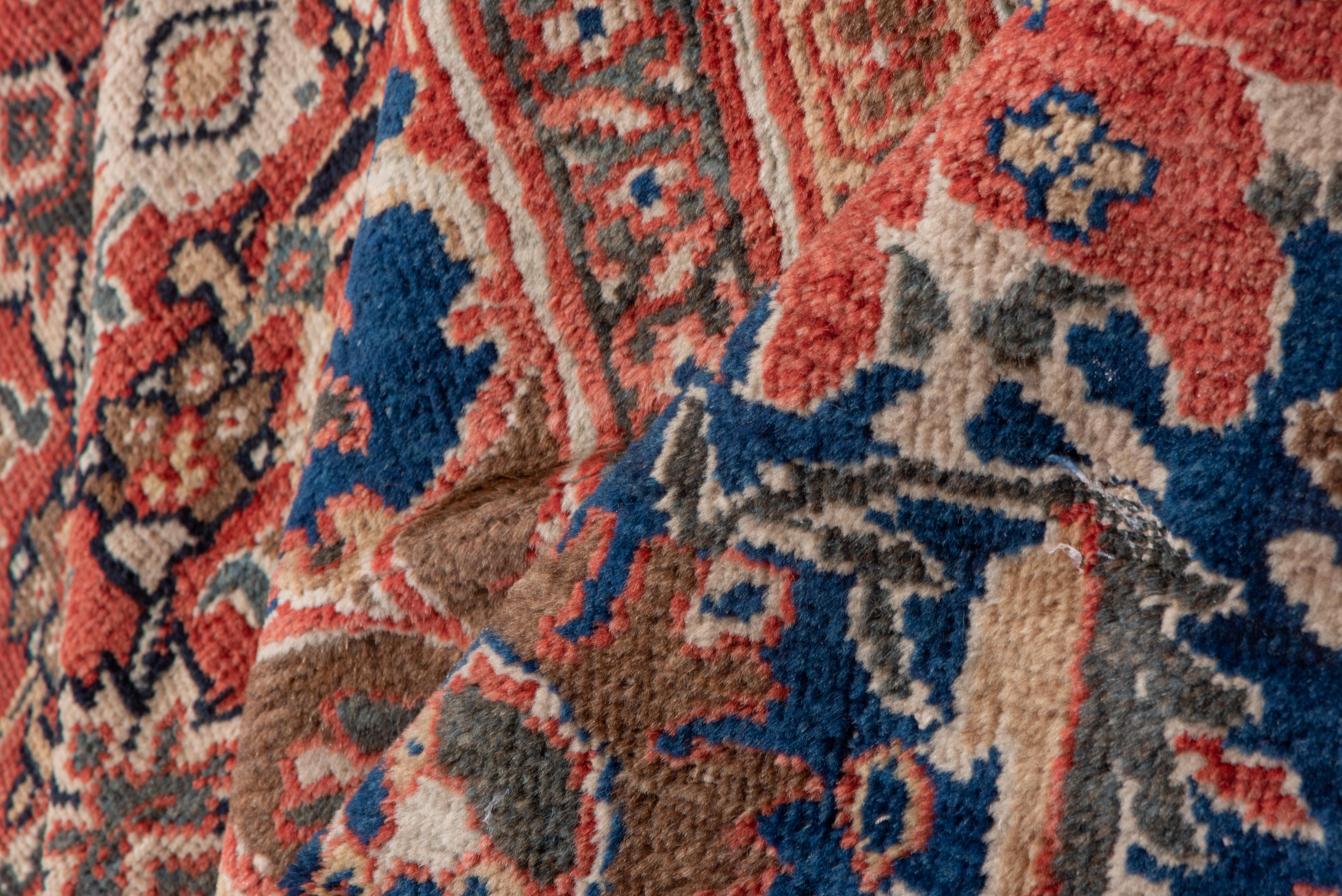This handsome very large west Persian village carpet shows a carefully drawn smaller Herati all-over pattern on a rust-red madder ground, fixed by a small royal blue ragged medallion and matching corners. Royal blue main border with two types of