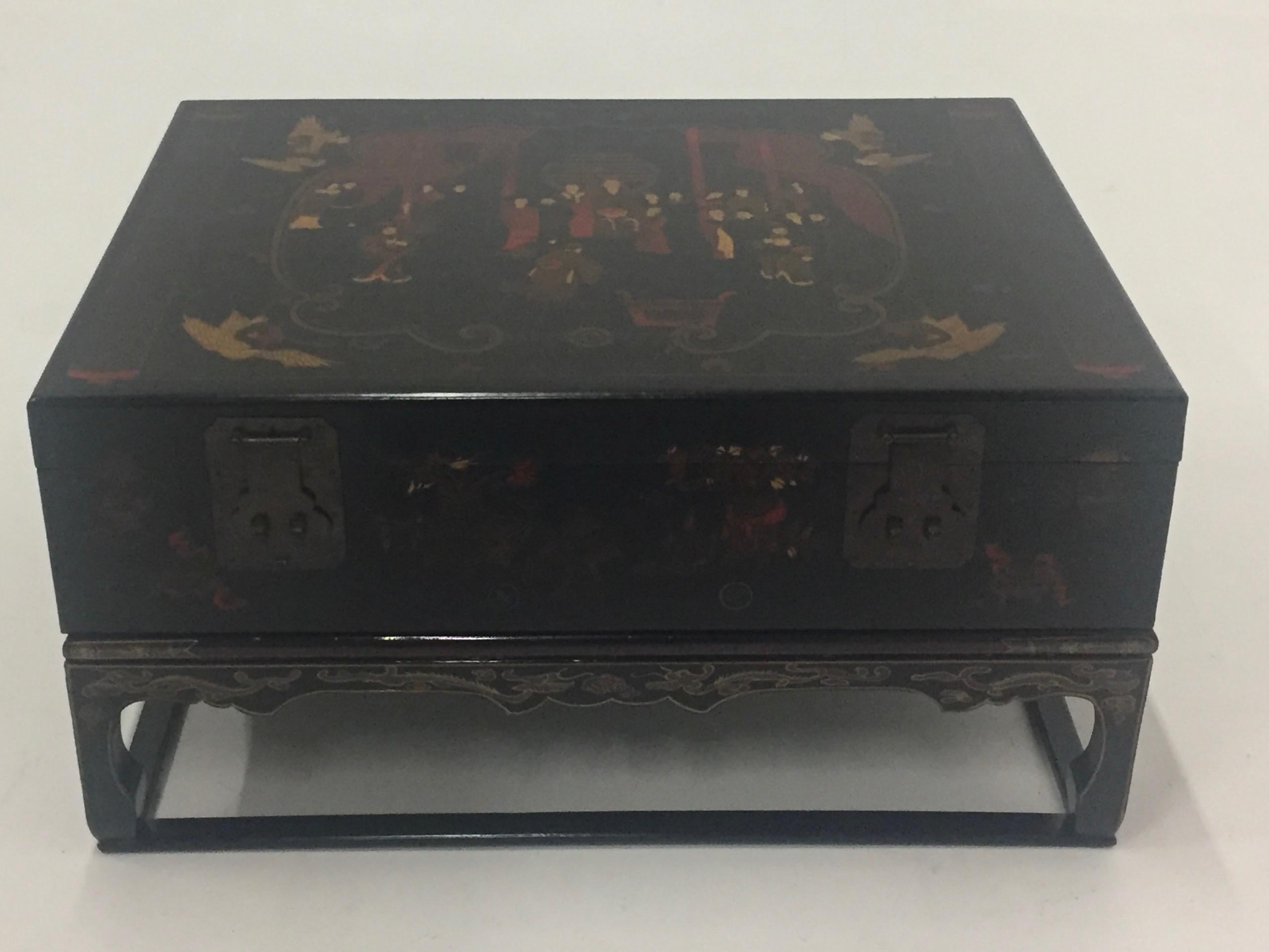 An intriguing chinoiserie coffee table in black, gold, and red having a beautifully decorated box with original hardware that sits on a custom ebonized Asian style stand. The box opens to reveal storage within and is lined with gorgeous decorative