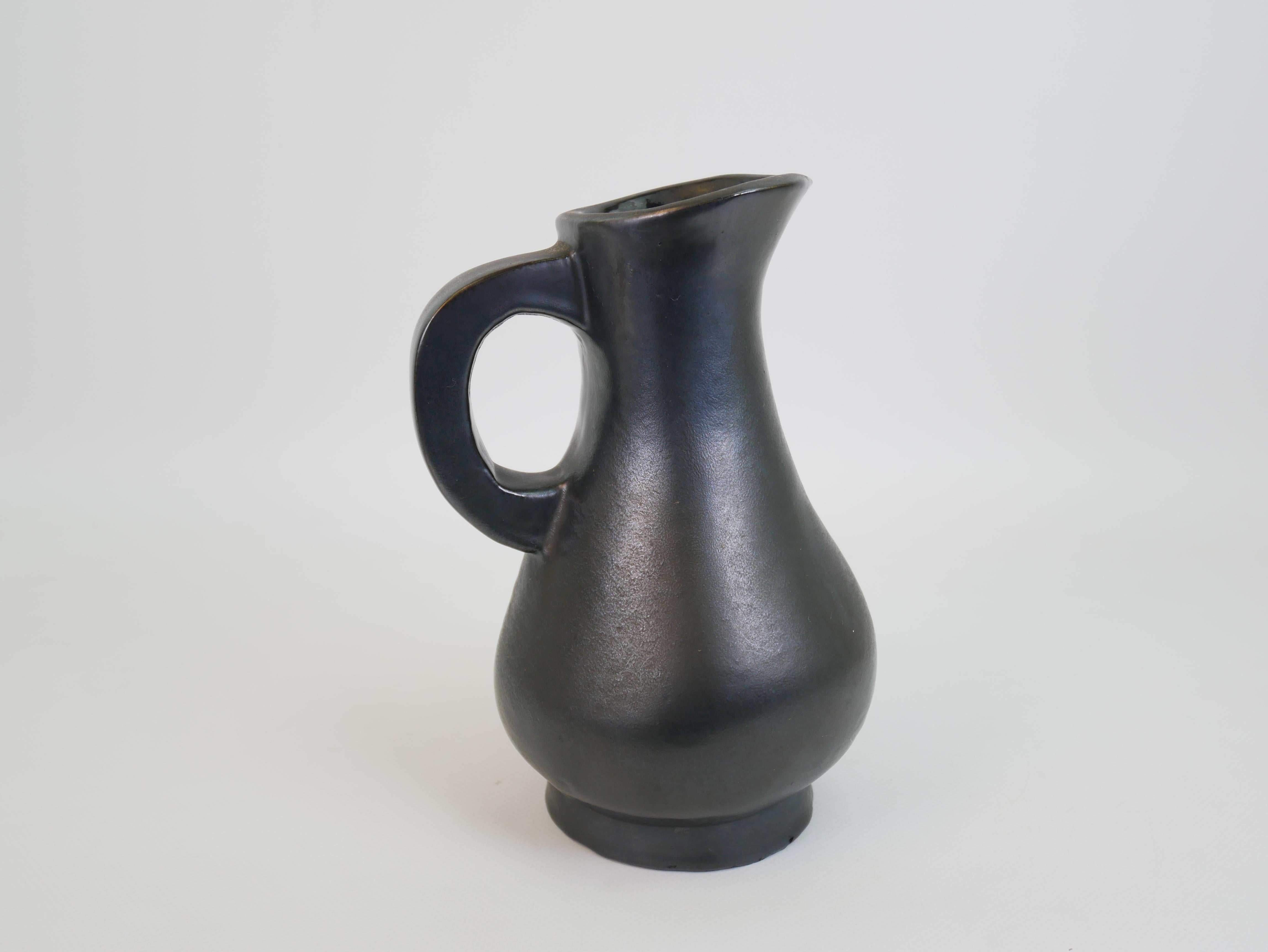 This elegant black-glazed ceramic pitcher embodies the spirit of mid-century French modernism, strongly reminiscent of the work of Georges Jouve. Its organic, curvaceous form features a gracefully flared spout and a rounded body that tapers toward a