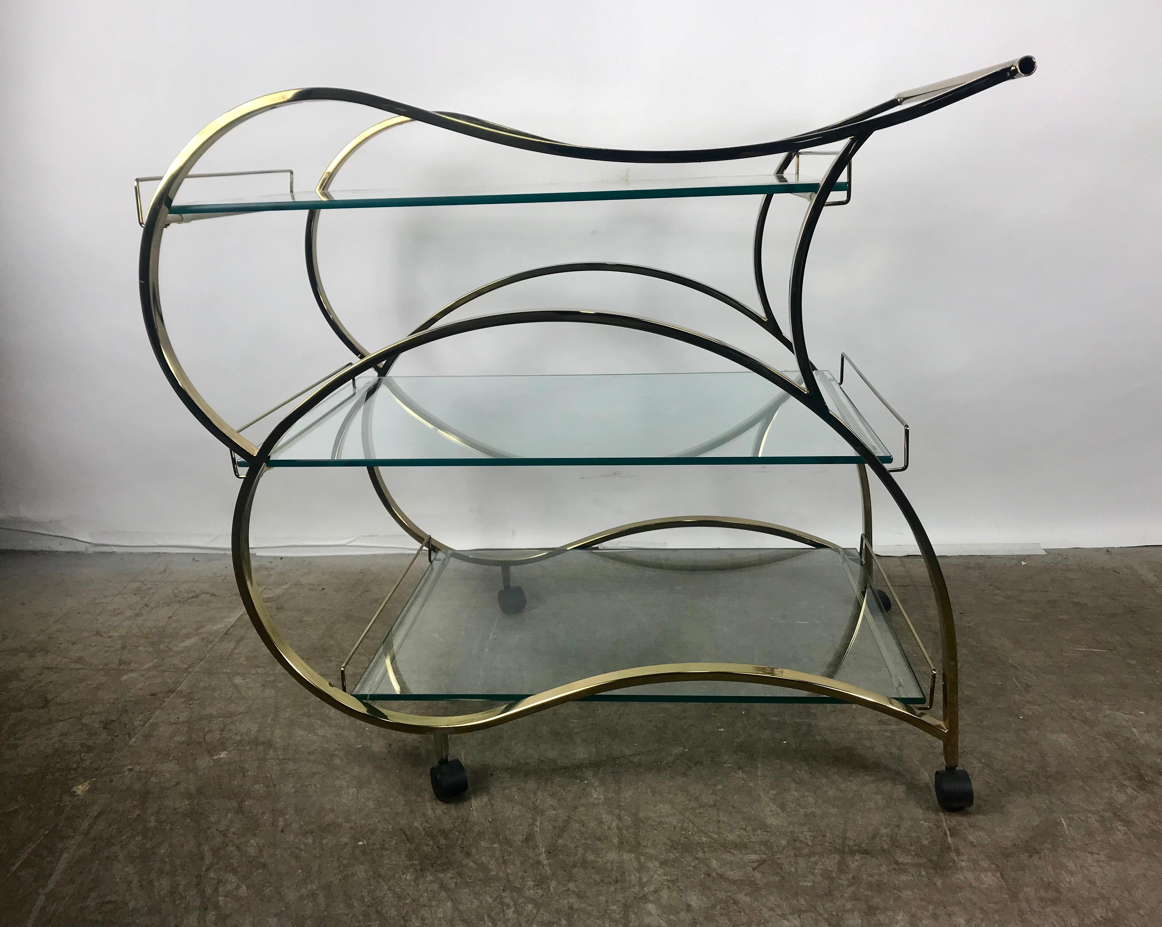 Stunning brass and glass modernist tea or bar cart, trolly seductive lines, streamline design, gleaming brass and 3 half inch glass with polished edges, makes a bold statement Show stopper! Hand delivery avail to New York City or anywhere en route