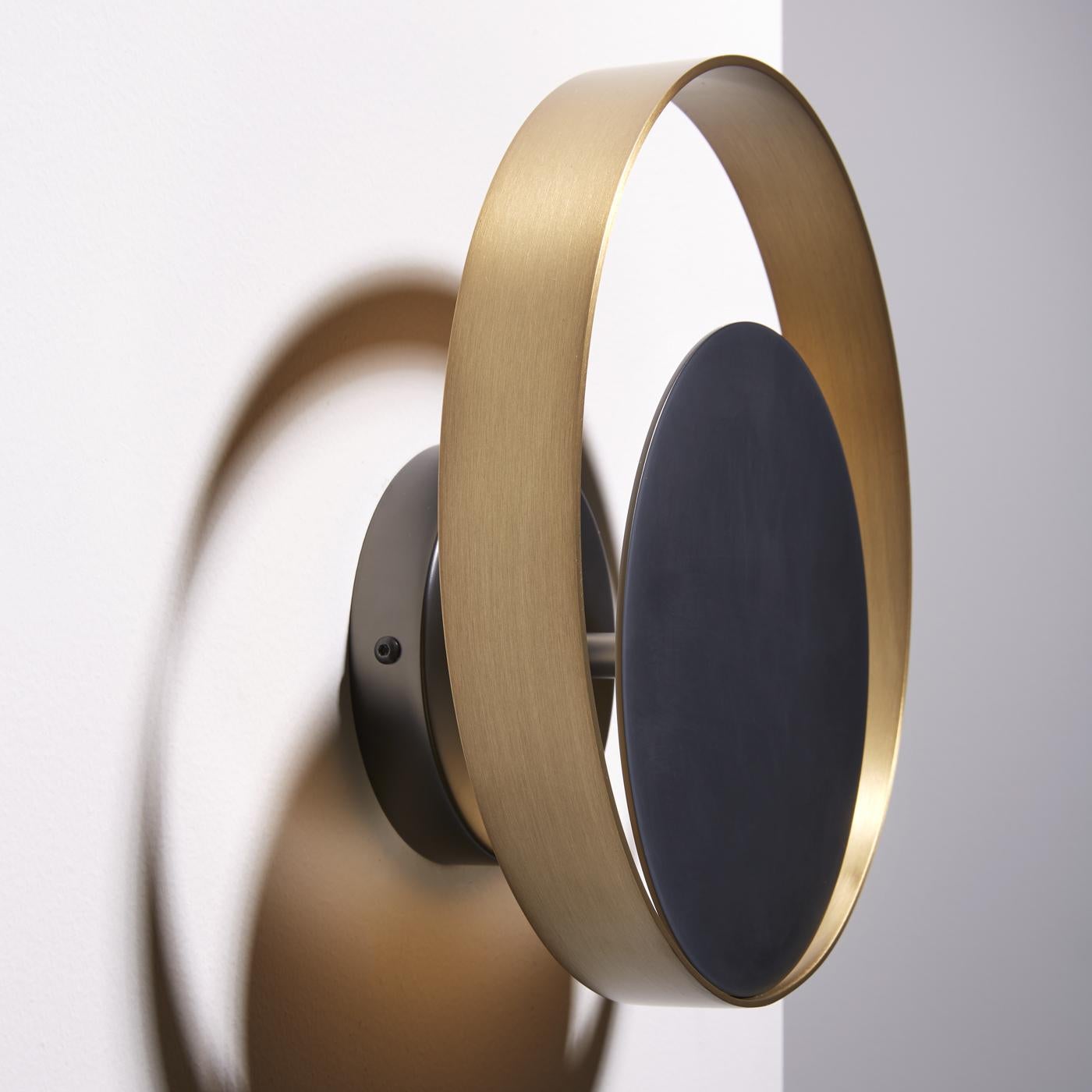 This sophisticated wall lamp projects an indirect light on the wall. It is comprised of a ring in natural brass surrounding a matte black nickel circle. This item is also available in other measures.