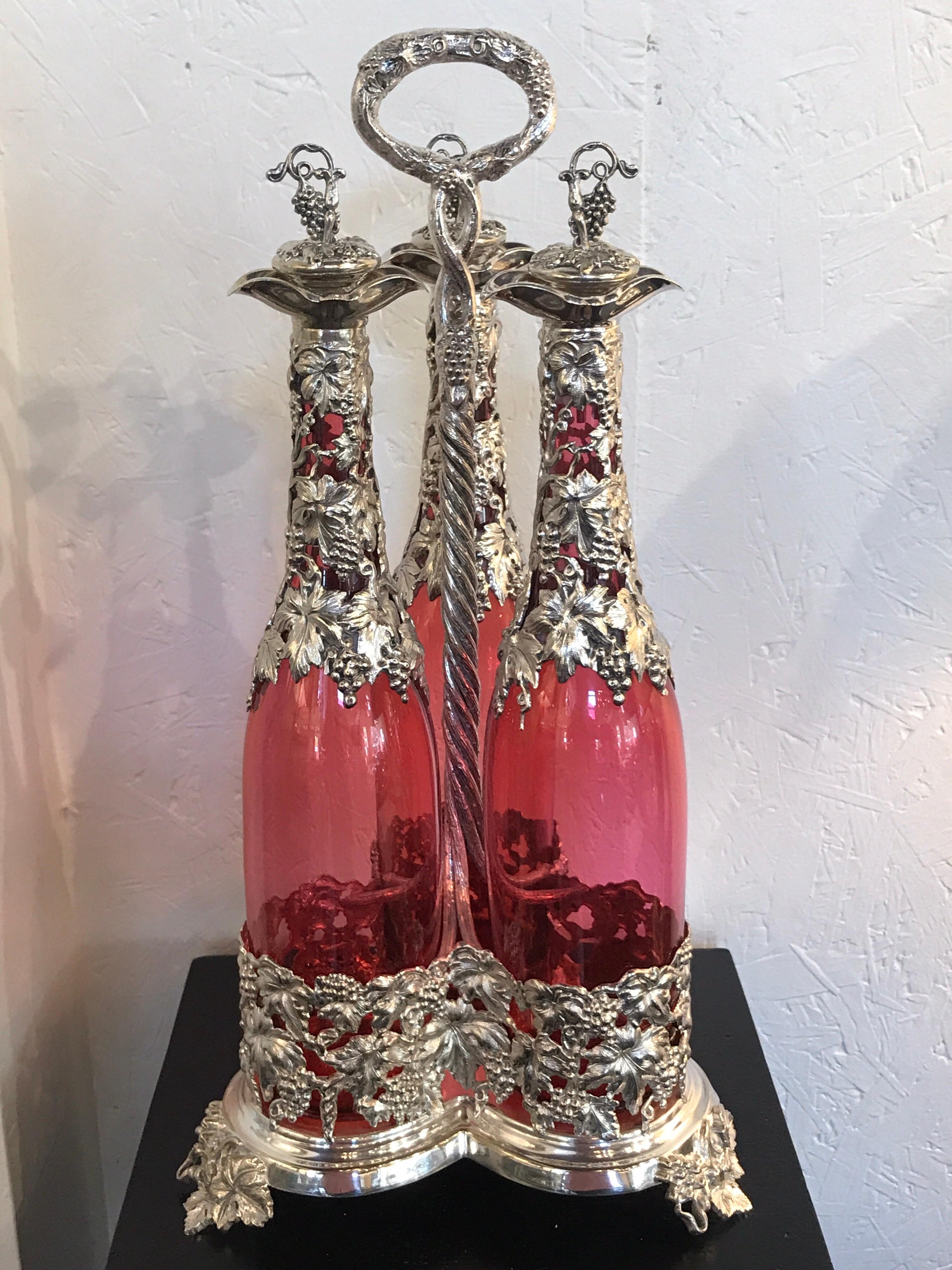 Stunning English silver plate and cranberry glass three-bottle Tauntless, with tall cranberry glass decanters dripping with applied silver work of grapes and vines. Fitted in an conforming, elaborate caddy, attributed to Elkington. Each bottle is