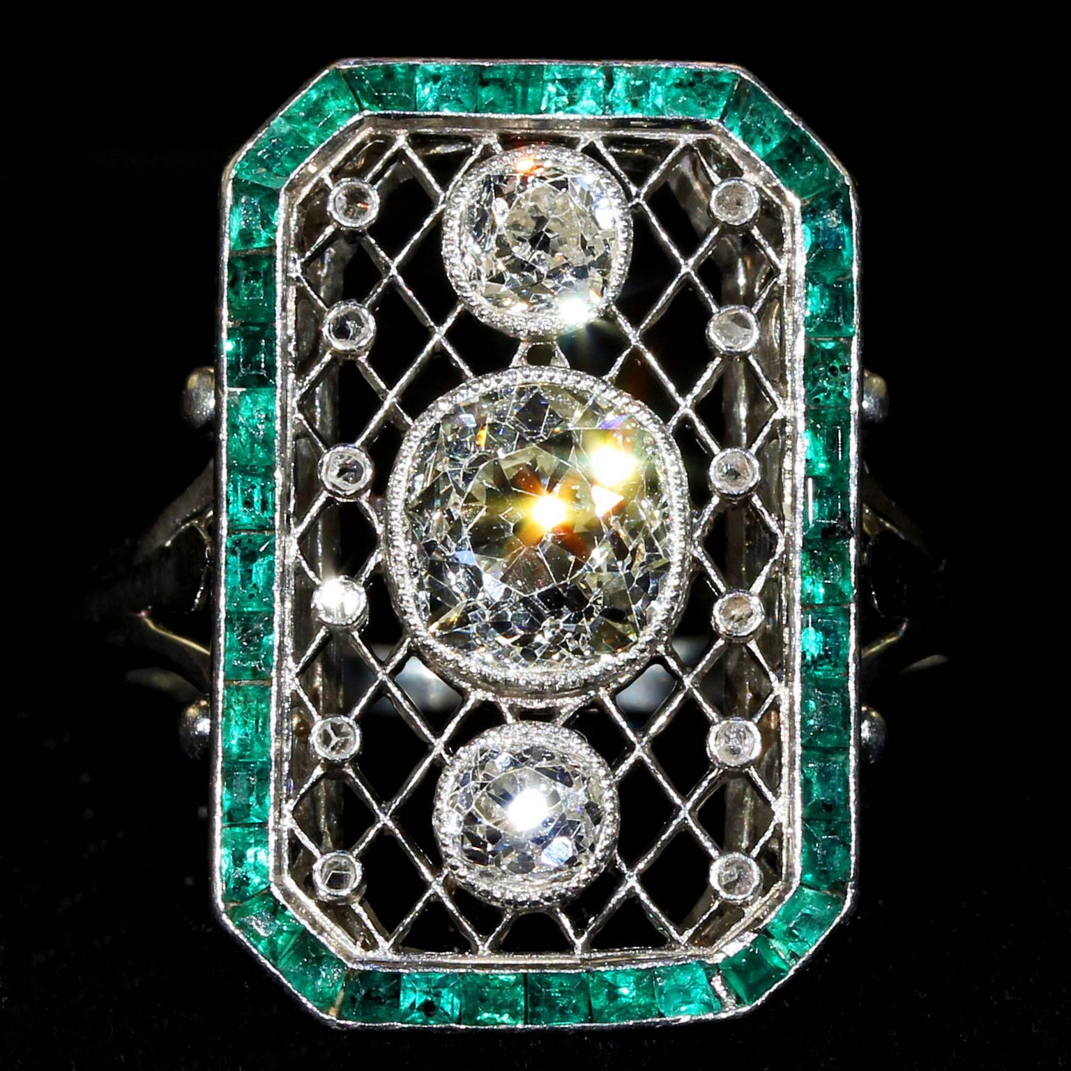 This circa 1910 French ring was hand crafted in Platinum with a rectangular low profile gallery. Three large center diamonds surrounded by a Platinum lattice work and a boarder of calibre cut natural Emeralds. Accenting the lattice work are two rows
