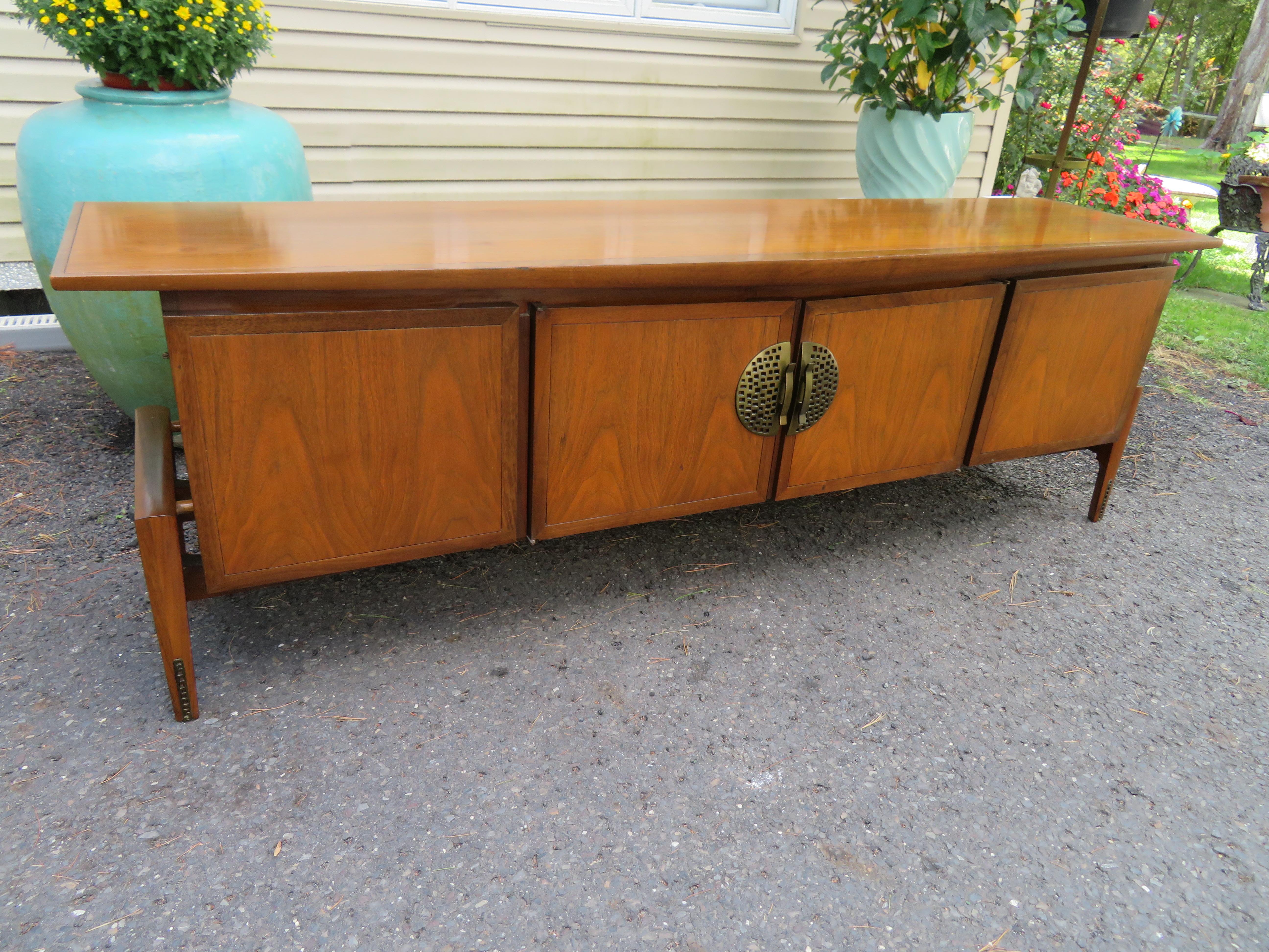 Stunning Helen Hobey Baker long low credenza Dresser with an Asian infusion. This sleek low walnut credenza has 2 open spaces on either side and 2 drawers hidden behind central doors. We love the interesting pierced large medallion hardware and the