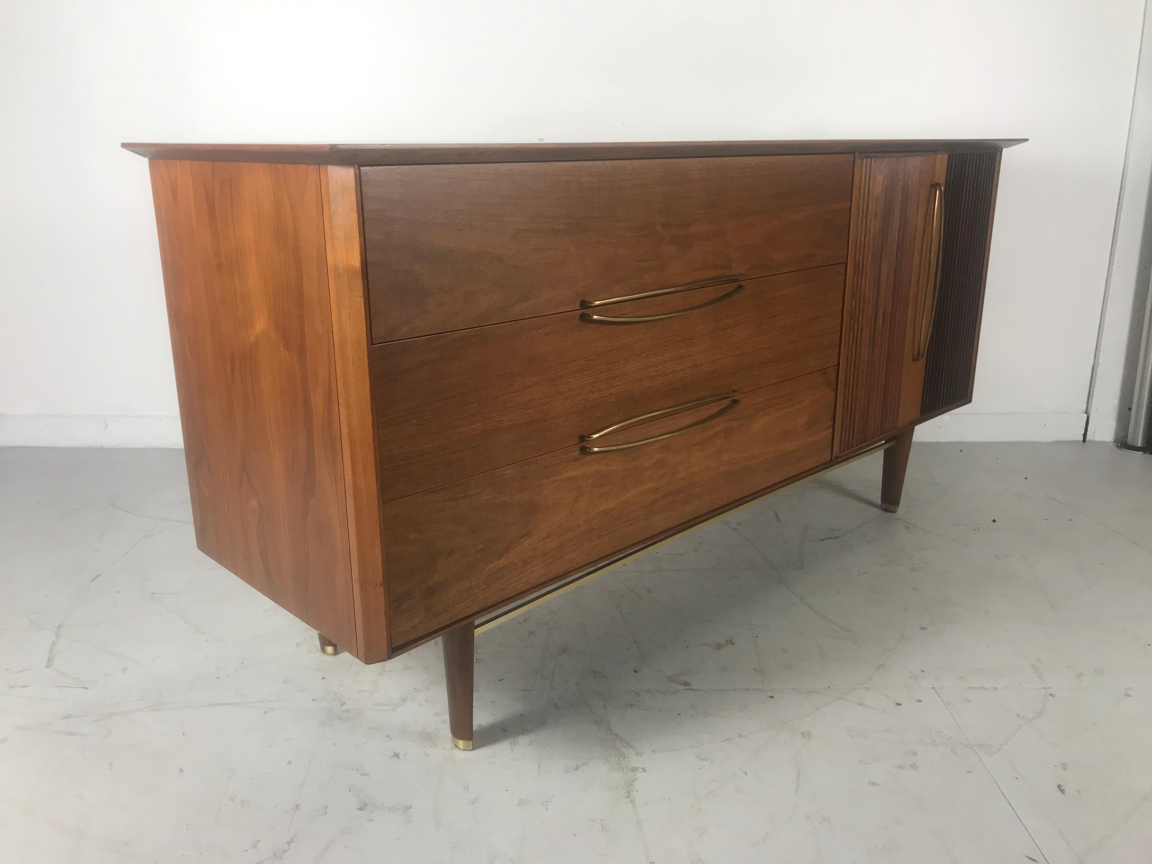 Stunning modernist walnut and brass dresser by Helen Hobey Baker. Superior quality and construction. Wonderful stylized brass hand pulls, three generous divided drawers (left), two doors with three drawers (right) Beautiful figured wood graining.