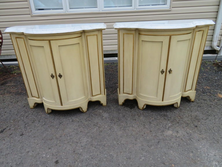 Lovely pair of Baker demilune console tables. They have a uniquely shaped marble top, over 2 cylindrical doors, raised on bracket feet. We love the original light yellow painted finish with the original brass hardware-has the original key. The