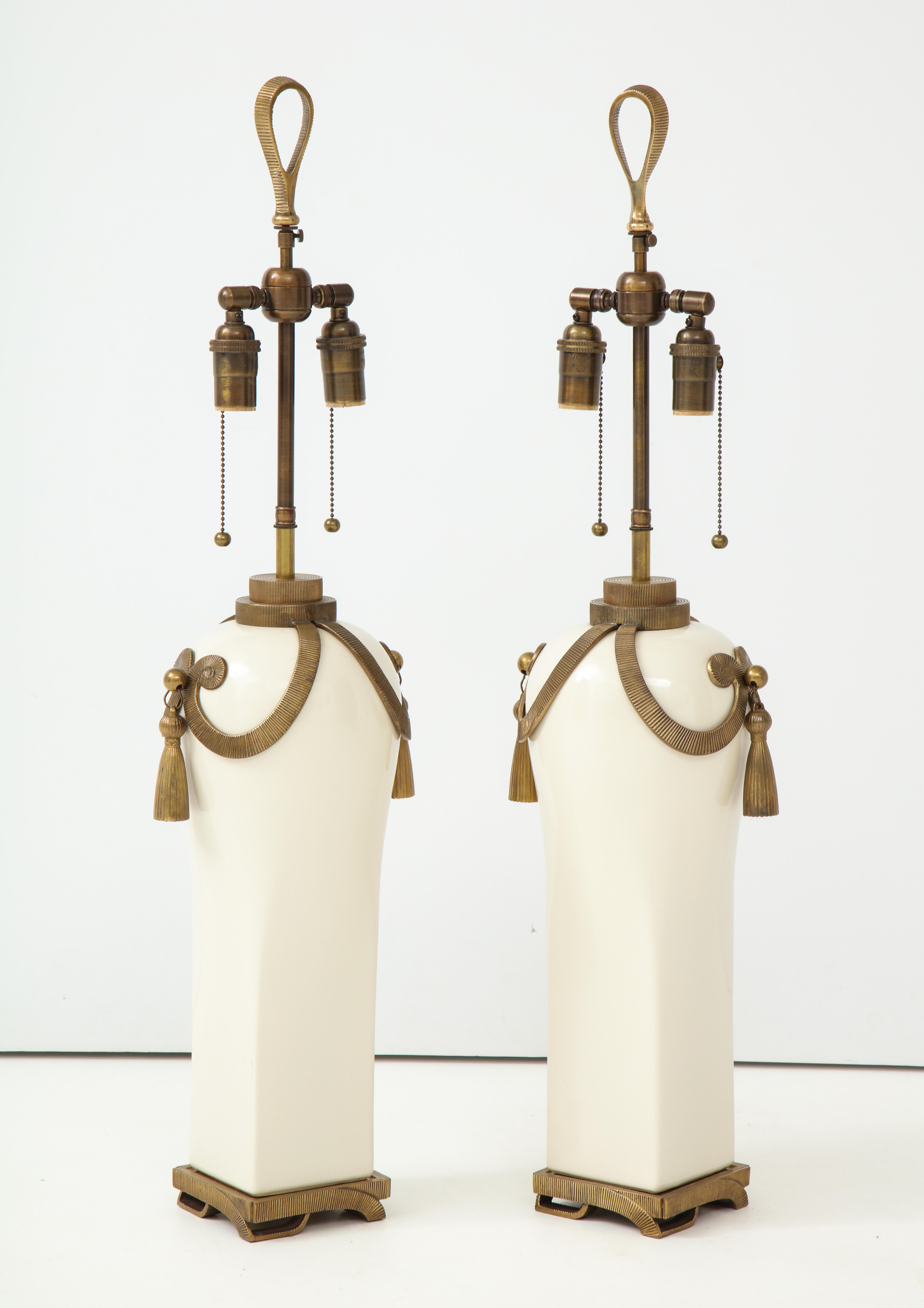 Pair of large ceramic lamps elaborately decorated with brass fittings
in the Art Deco style by Chapman.
The Ivory glazed ceramic lamps are decorated with a grooved brass patterned trim which is followed throughout with matching heavy tassels that