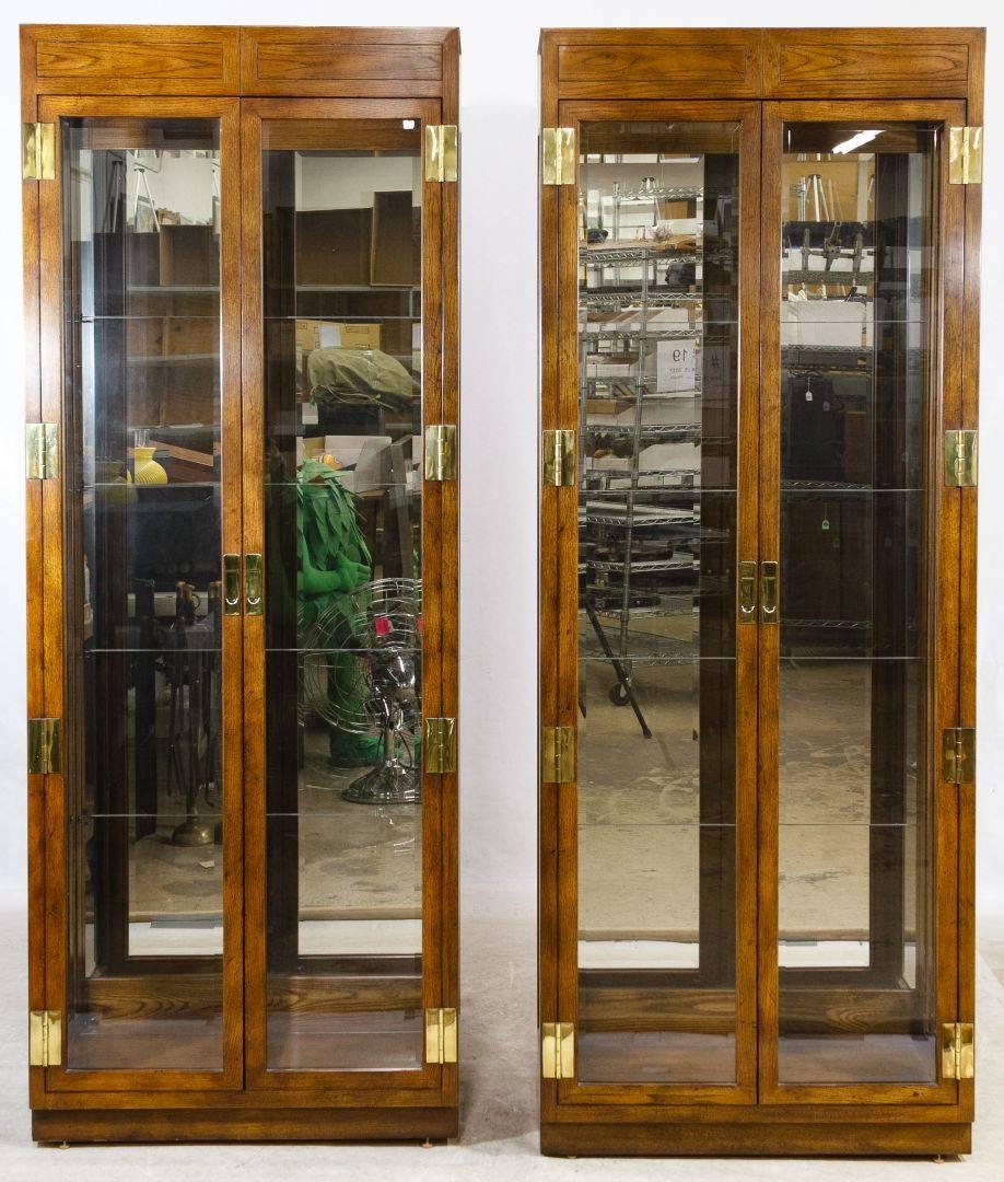 A stunning pair of Campaign style curio display cabinets. The cabinets feature two beveled glass doors which open to an interior with a mirrored back. There are four thick glass shelfs in each and two lights in the top. Heavy brass hardware adorns