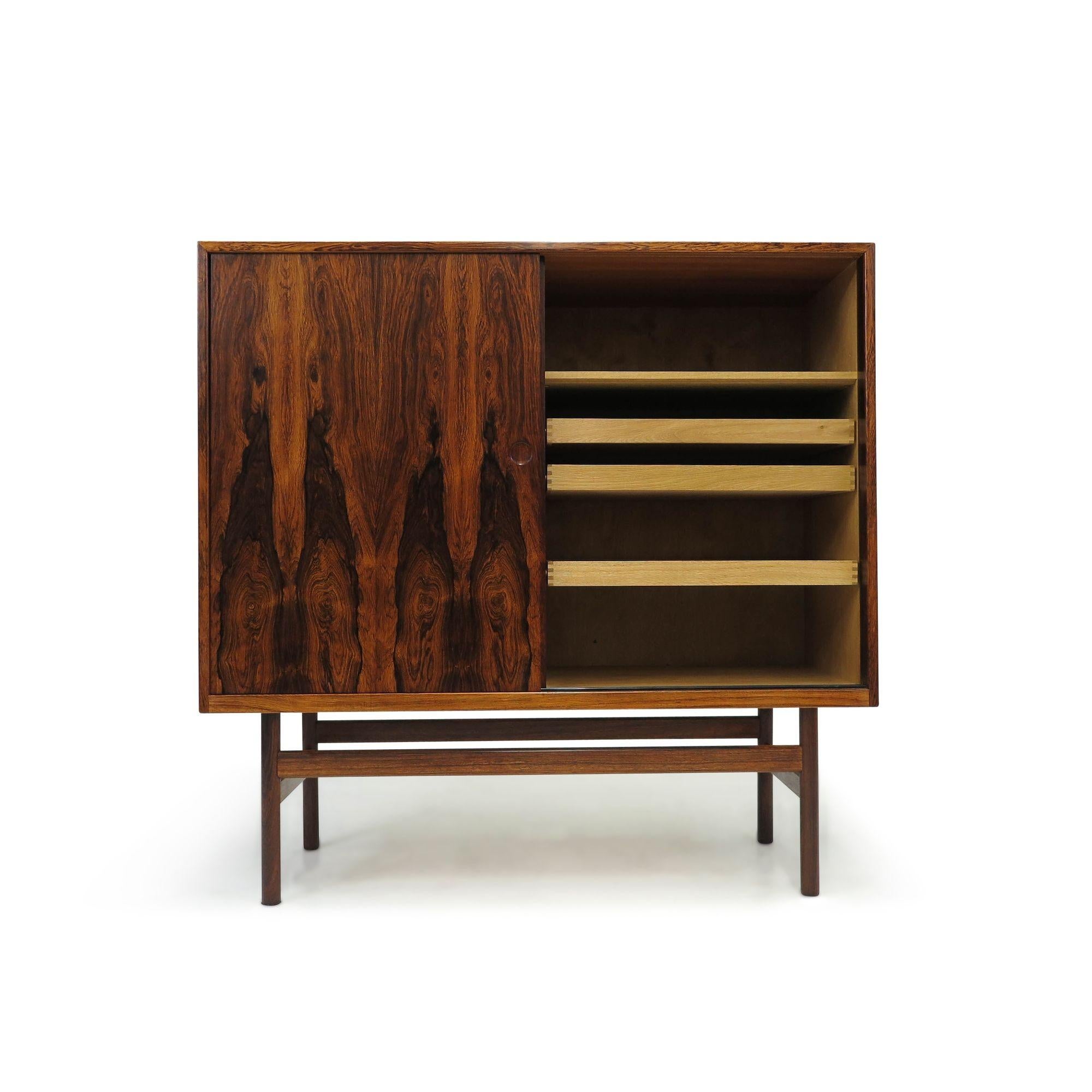 Danish cabinet with stunning book-matched Brazilian rosewood, showcasing dynamic grain patterns on the sliding doors front. The cabinet doors open to reveal a white oak interior with silverware drawers and adjustable shelves. The cabinet stands