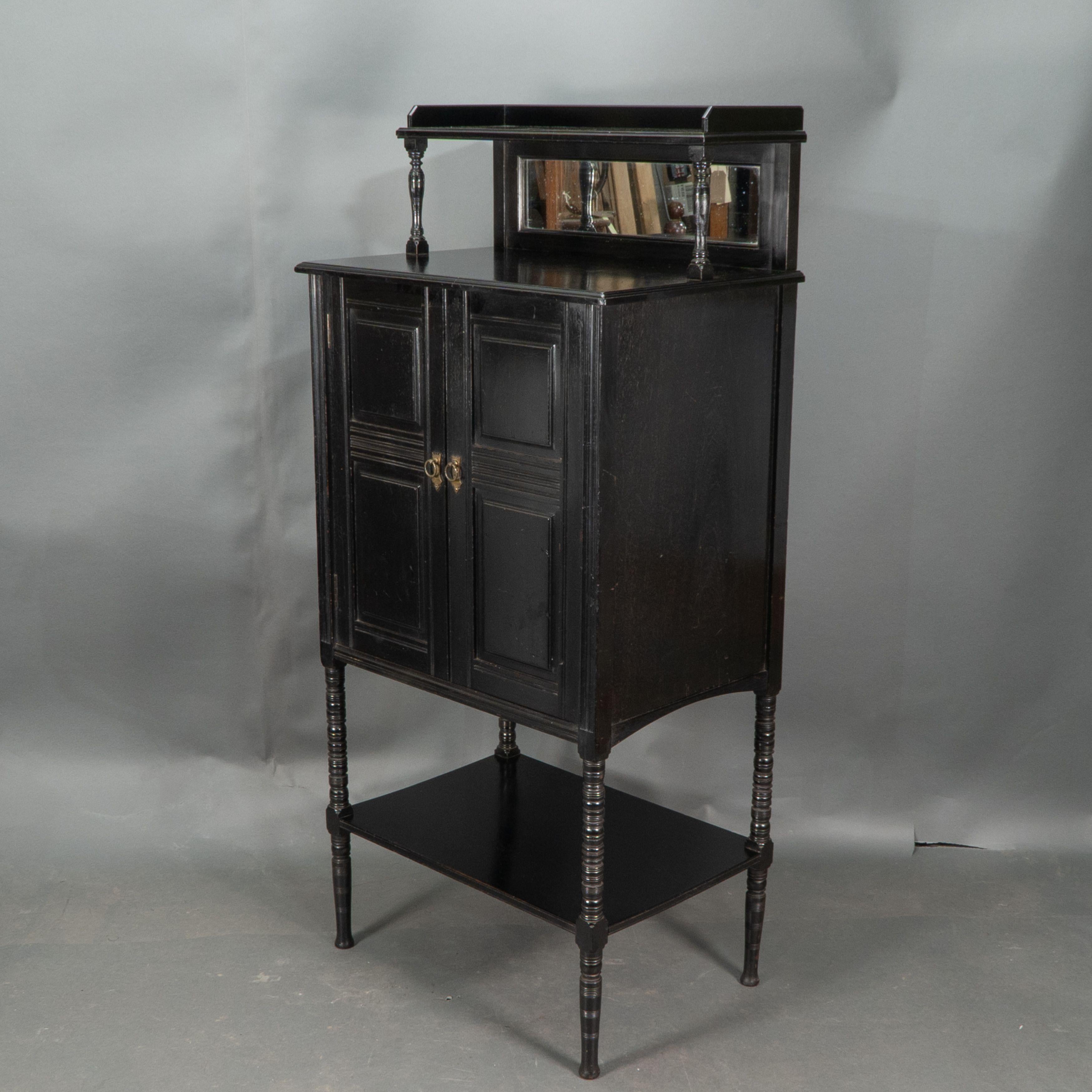 Style of E W Godwin. An Aesthetic Movement ebonized cabinet. The upper shelf with a 3/4 gallery on turned supports with a bevelled mirror below and display area in front. The two door cupboard with four fielded panels centred by tramline decoration