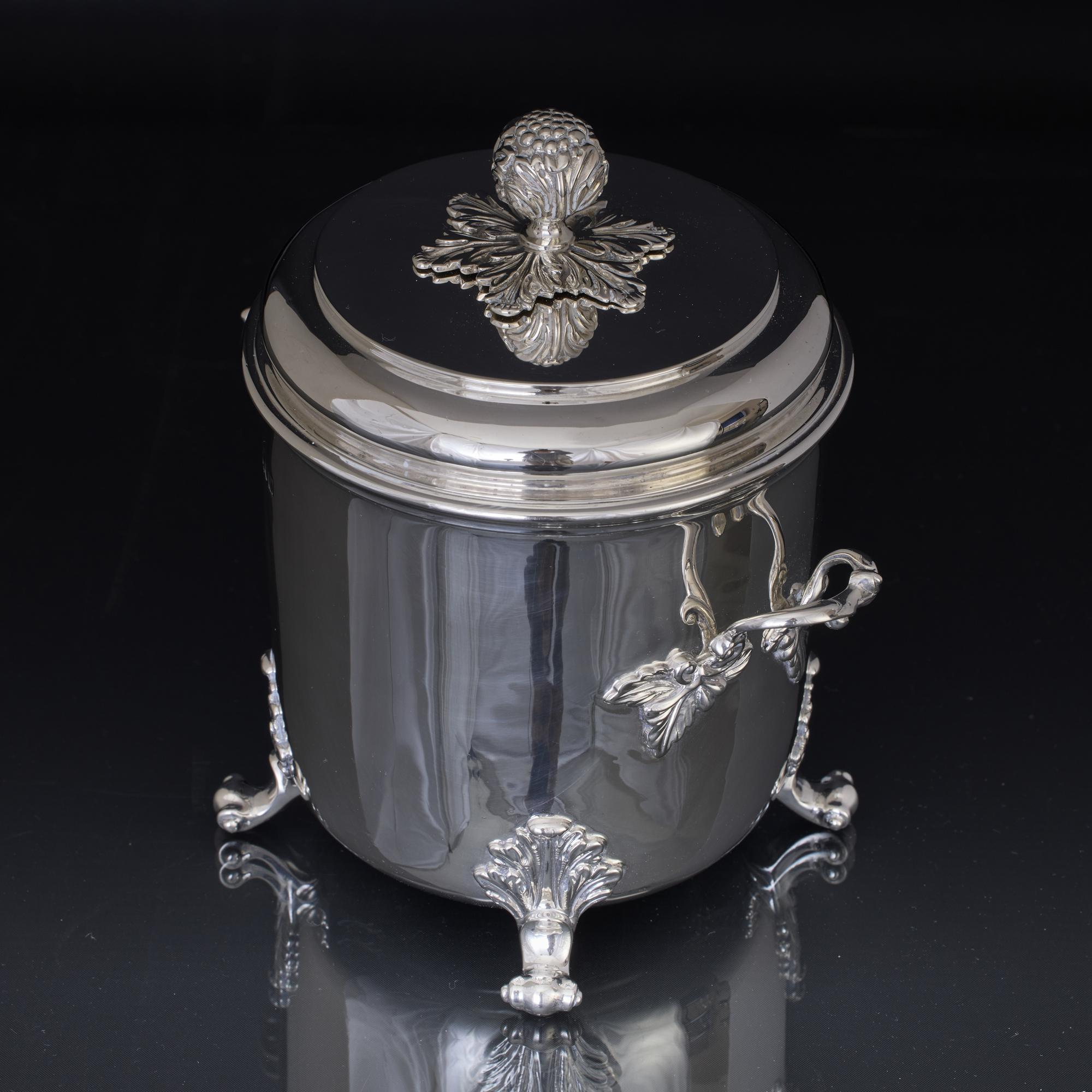 Importante recipiente cilíndrico de plata antigua con dos asas, tapa abatible y montado sobre cuatro soportes de fundición con volutas y hojas de acanto. Las dos asas de fundición se ajustan a la caja con cierres decorativos de hojas, mientras que