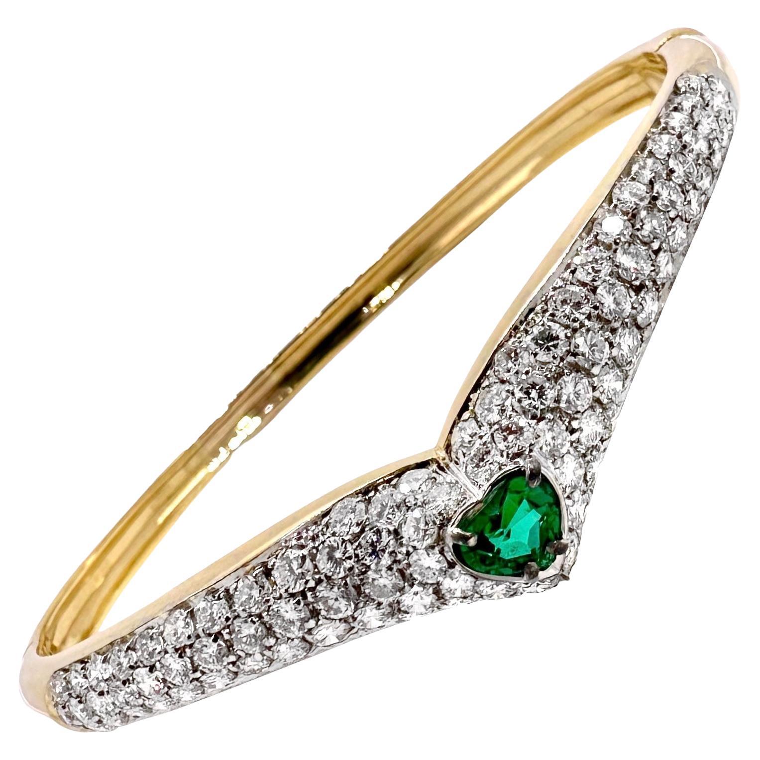 This stylish and dazzling 18k yellow gold hinged bangle bracelet is unmistakably a product of the 1970' or 1980's. Set at the center of a bombe chevron form plate, is an absolutely vivid heart shape natural emerald. Framing this small gem are long