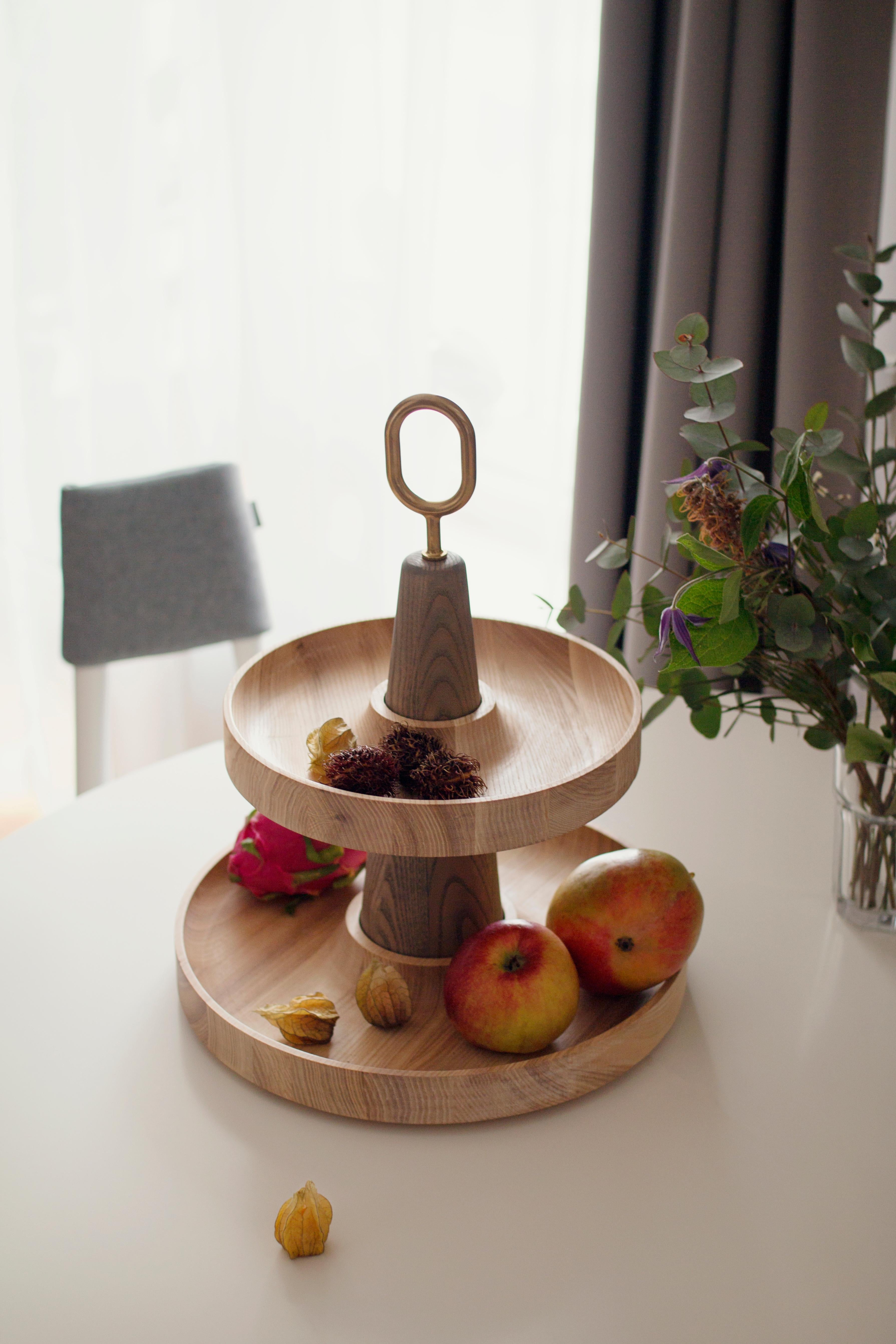 The tiered serving stand Babel is a composition of two scooped wooden plates and a central pole combined with a solid brass handle. All three parts are made with precision from solid ash, and the plates are oiled with oil which is certified for