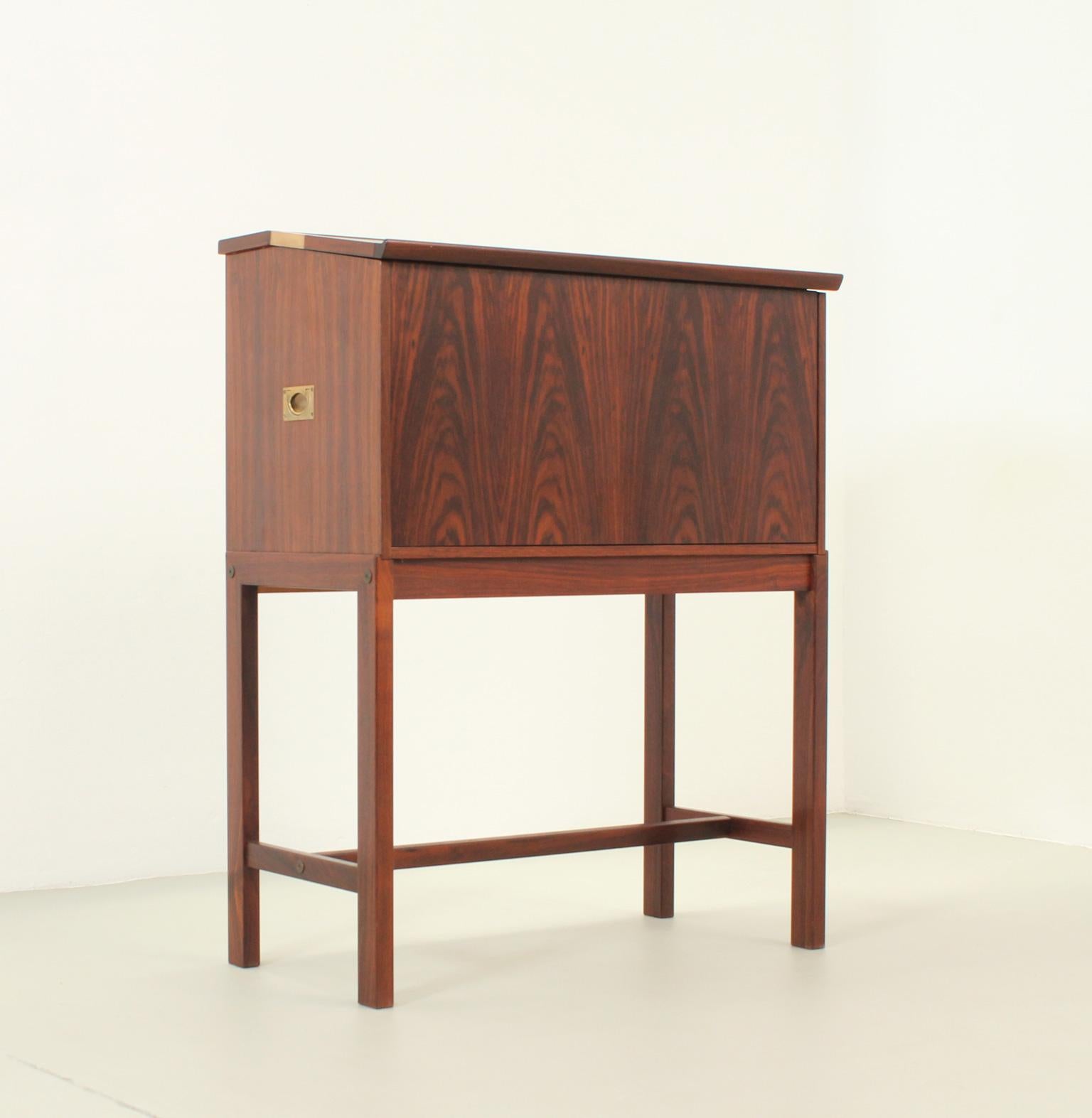 Standing bar cabinet produced by Dyrlund, Denmark, 1960's. Stylish design with the front part that folds down to create a serving surface, and interior with a hole for bottles. Hardwood, brass and leather. 

Measurements when it is open 79 cm.