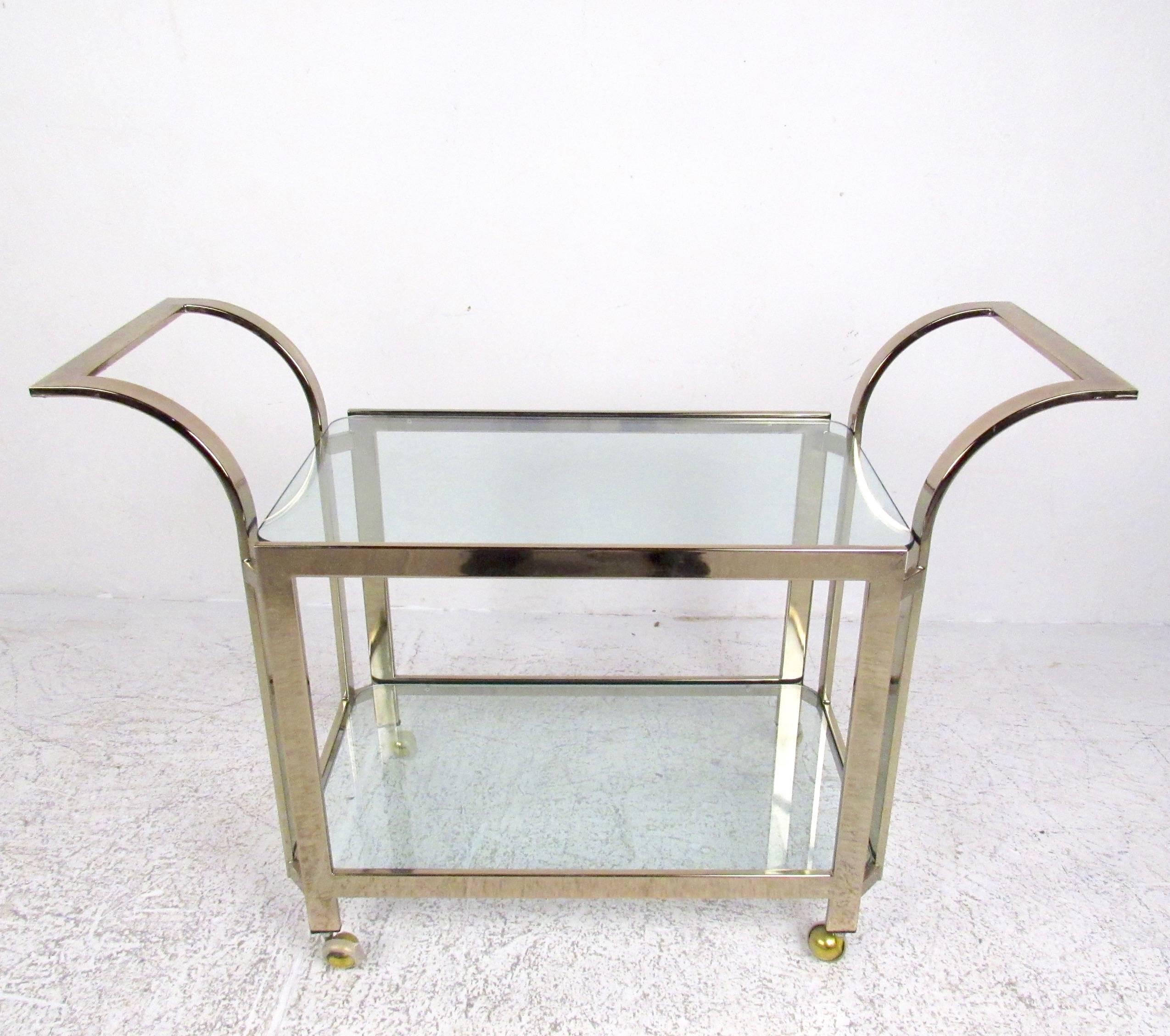 This vintage modern serving cart features two-tiered glass shelves for bottle or glassware storage. An ideal shop display or bar service cart, the shapely modern design and vintage brass finish add to the Mid-Century Modern appeal of this versatile