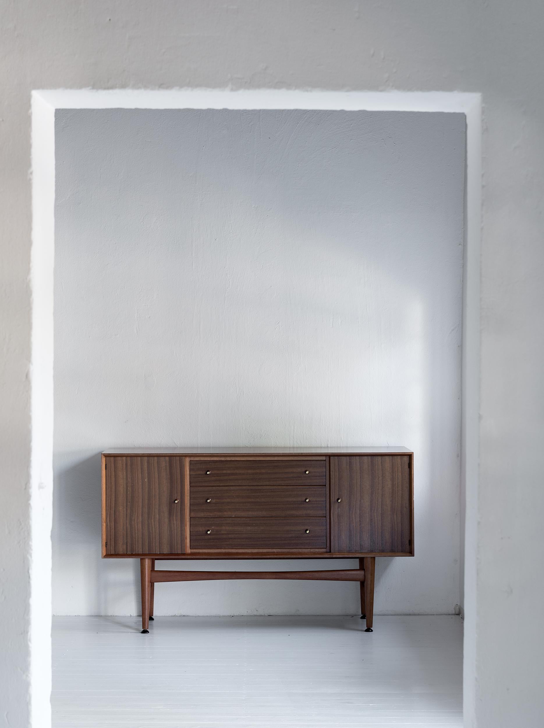 Beautiful rare sideboard by Gordon Russell in teak. Two spacious compartments with adjustable shelving flank three drawers. Superbly finished with the signature teak and brass trumpet style handles. In great condition and very chic. 

Gordon