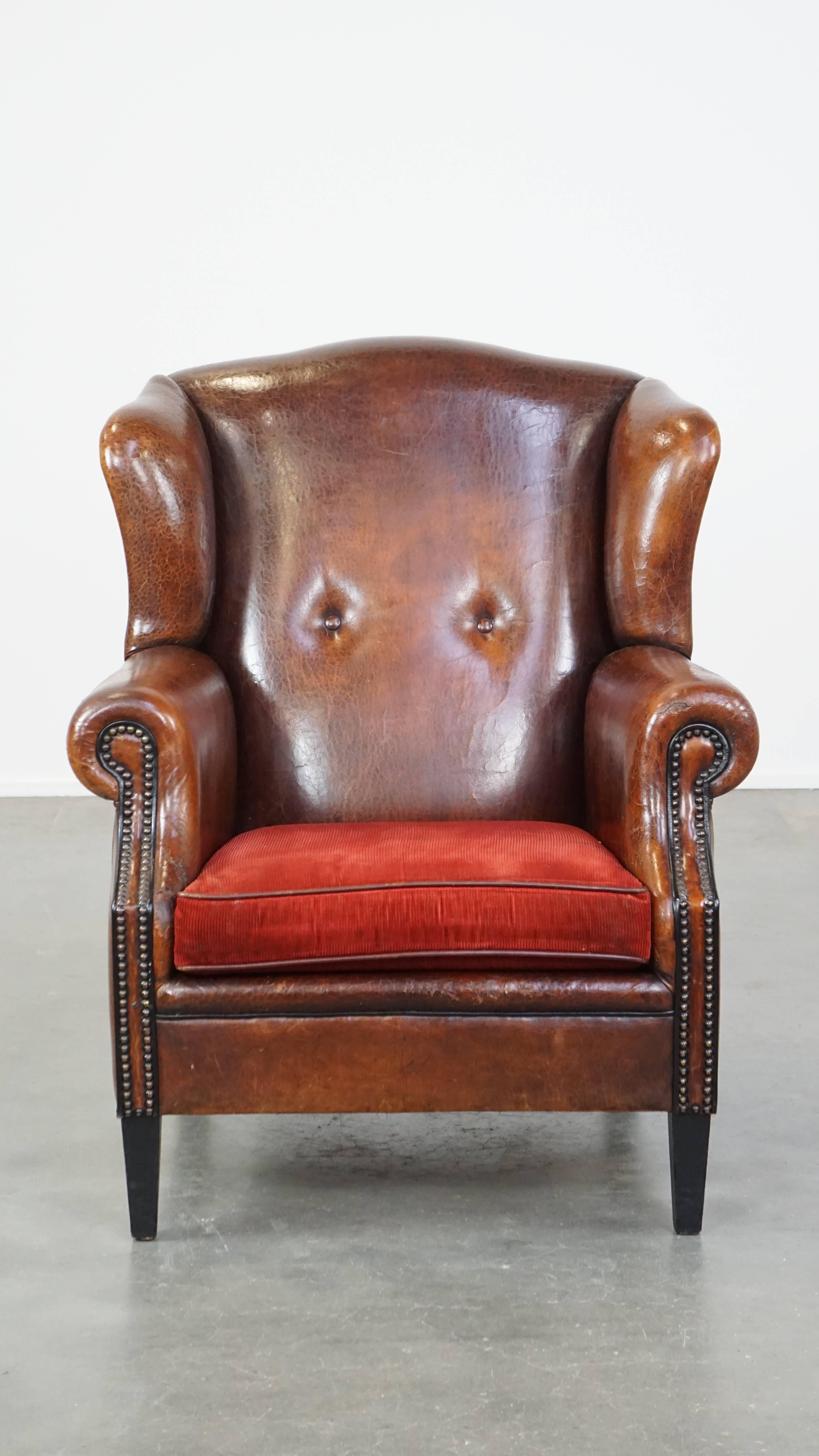 This sheepskin armchair, with its deep colors, red corduroy seat cushion, and nailhead trim, exudes a luxurious and refined character. It is beautifully finished and has developed a stunning patina. The armchair features a loose seat cushion and