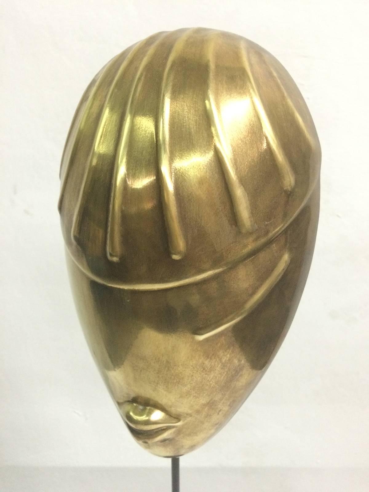 This sculpture is a stylized representation of a female face in brass. The oval face is finished with lines representing hair or a hat, one eye and lips. The piece is mounted on a piece of rod and a heavy steel base finished in black. Dimensions of
