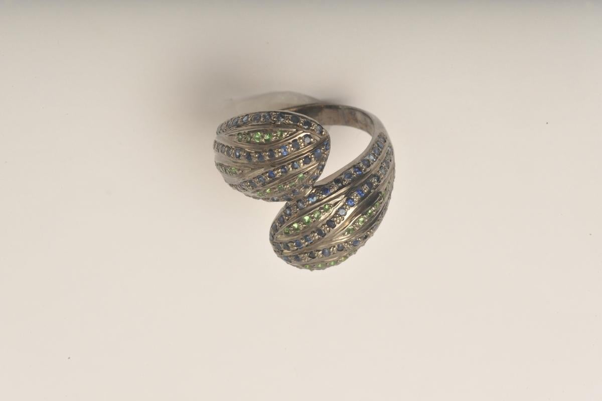 A stylized, double snake head ring with faceted, pave`-set blue sapphires and tsavorite stones gracing the top of their heads.  Set in an oxidized sterling silver.    Ring size is 7.75.