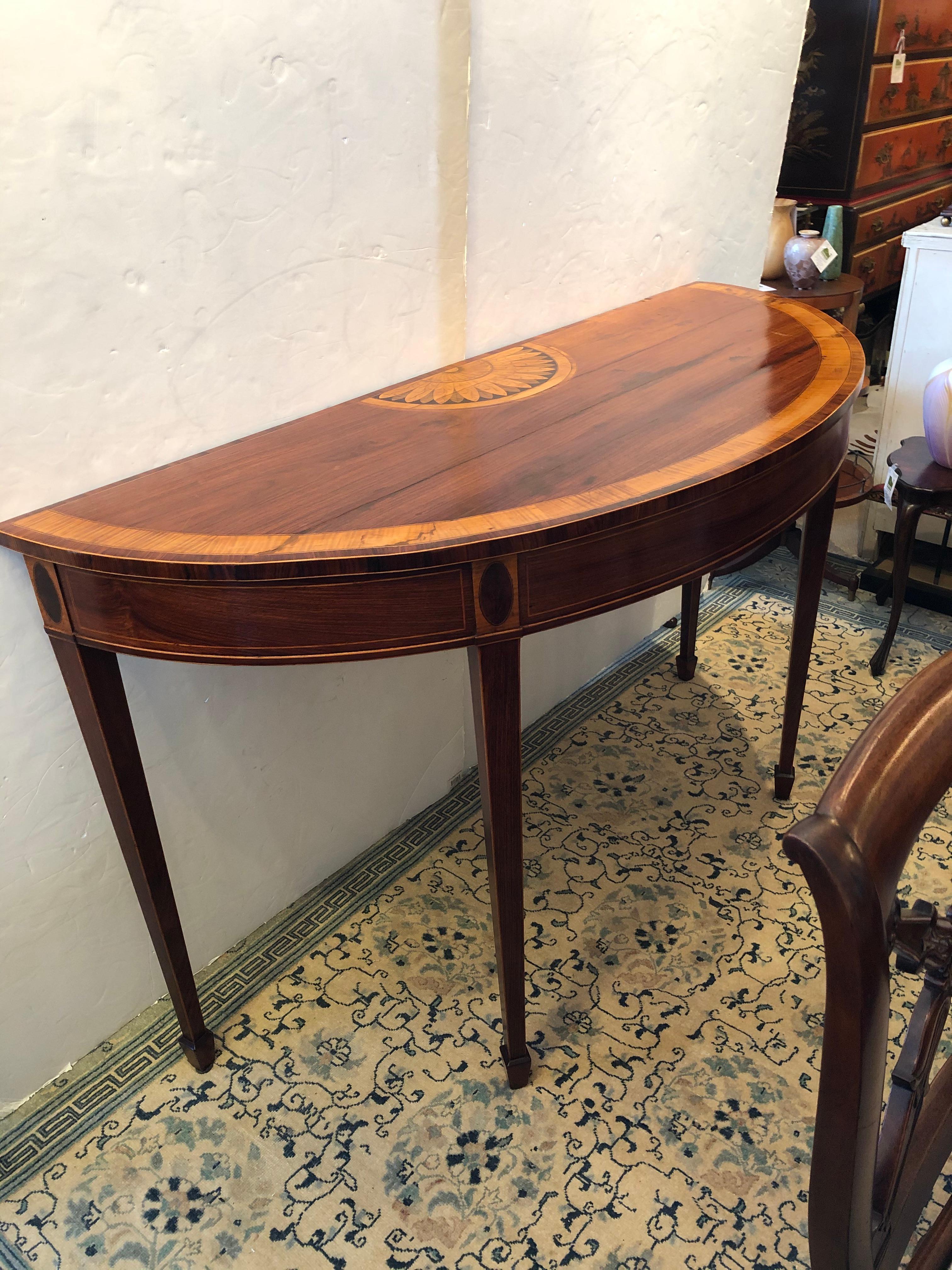 Incredible large and tall stately George III mahogany and satinwood inlaid demilune console having gorgeous half moon sunburst medallion at the back and other decorative inlaid flourishes, supported by elegant tapered legs.