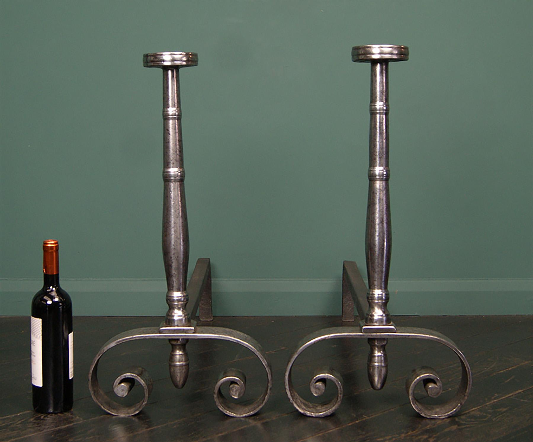 A substantial pair of wrought fireplace andirons with shaped collared standards surmounted with a disk.  The upright is supported on scrolled feet with a central drop finial, the rear log-supports can be adjusted to fit the fire opening. 