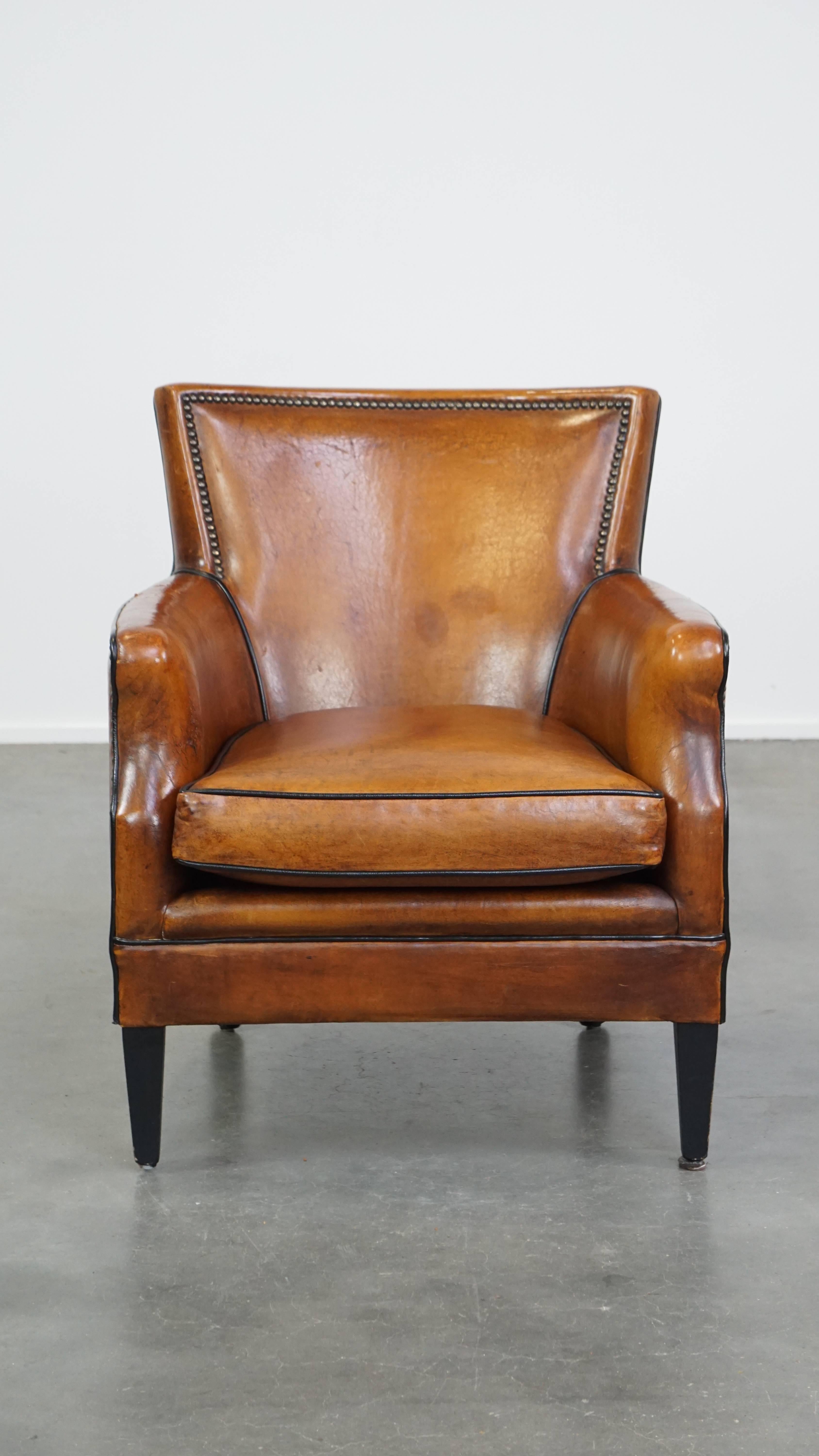 Questa poltrona in pelle di pecora color cognac ne è un esempio perfetto. Ha un design delizioso ed è rifinito con profili neri e chiodi decorativi, che gli conferiscono un aspetto raffinato e senza tempo. Questa poltrona è dotata di un cuscino di