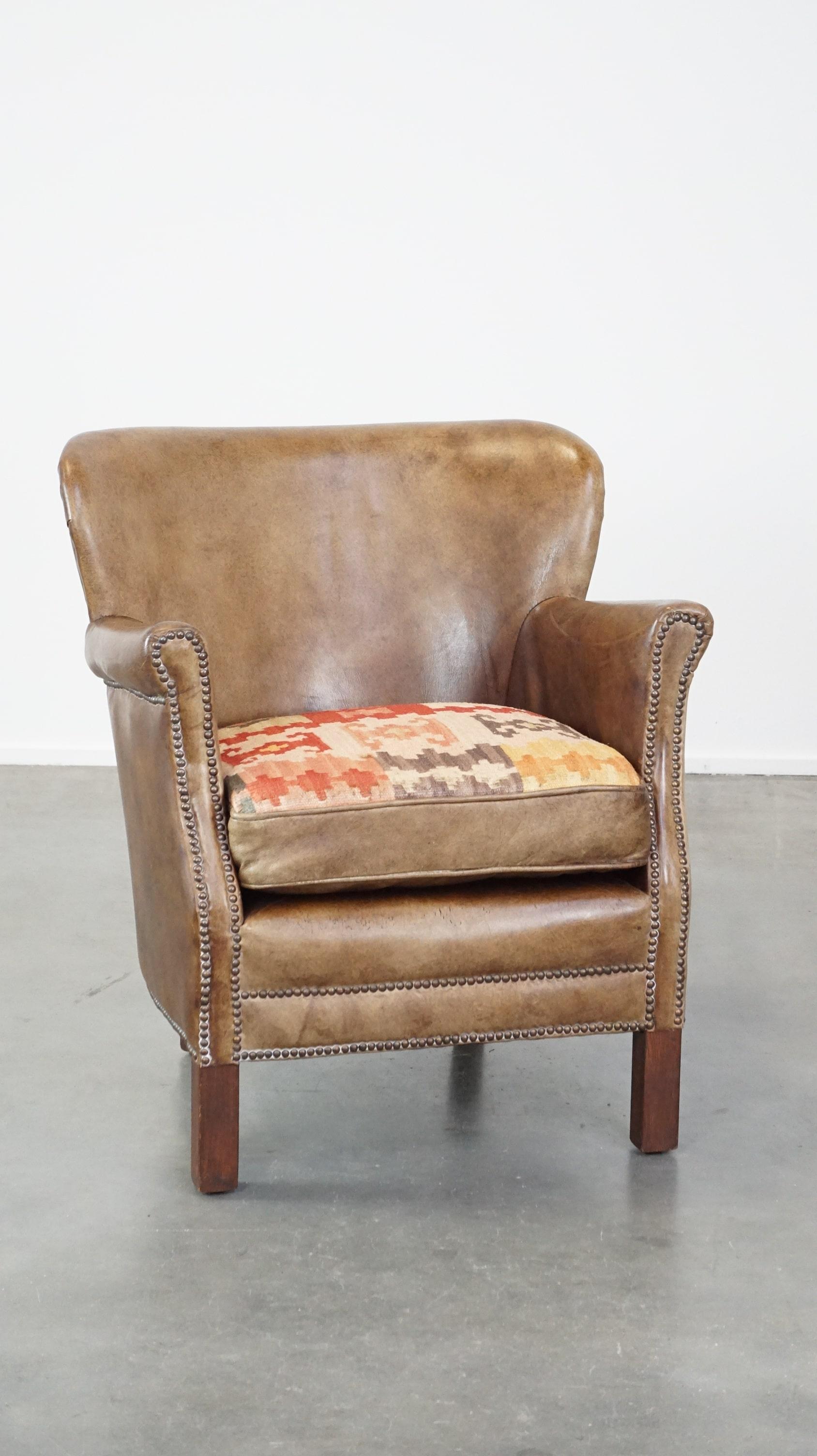 Ce fauteuil en cuir super charmant a un caractère distinctif. Il est légèrement décoloré d'un côté en raison de l'exposition au soleil et comporte un coussin d'assise amovible, caractéristique et réversible. Un côté du coussin est en cuir, tandis