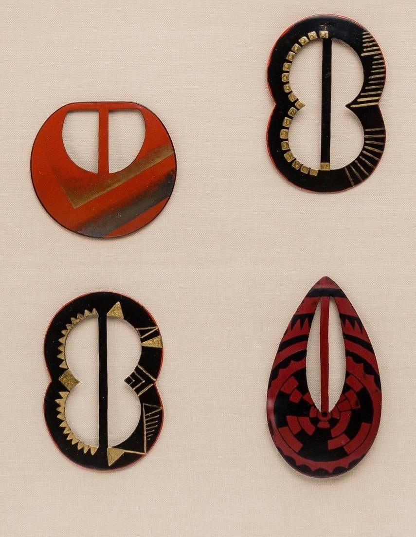 A set of four belt buckles with lacquered decoration. One of truncated circular shape with geometric gold and silver decoration on a coral background. The two other buckles are in the shape of an 8 with gold Africanist decoration on a black