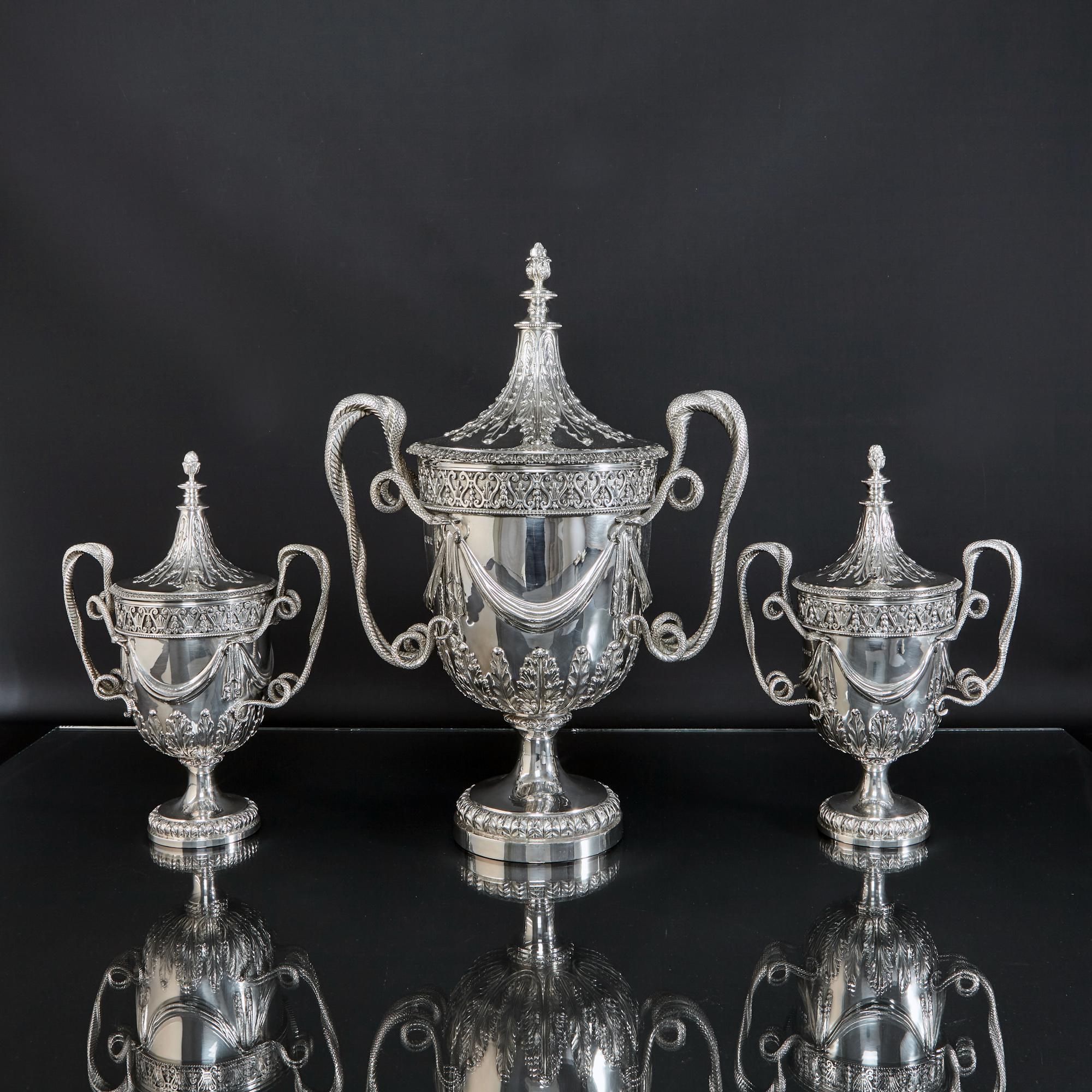A spectacular set of three silver trophy cups in the George III style, one larger and a pair of smaller. Each has a removable lid with hand-chased acanthus leaf detail, a decoration which is also found round the bodies, and cast finial. The handles