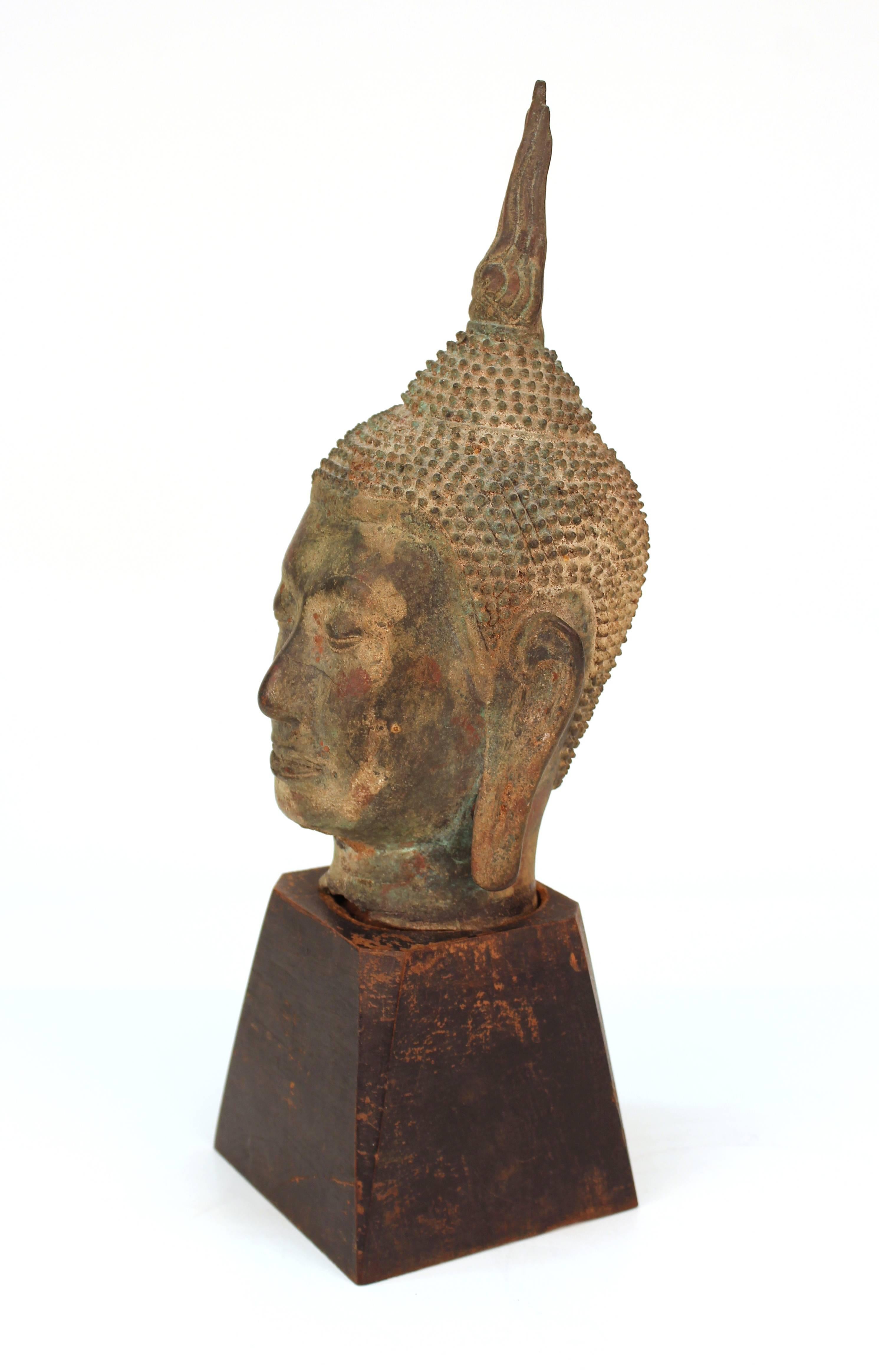 Sukhothai style Buddha head in bronze from Thailand. Crafted in bronze and mounted on a painted wooden base. The head can be removed from the stand. Wear includes natural discoloration due to age and uneven neckline. There is some wear to the paint