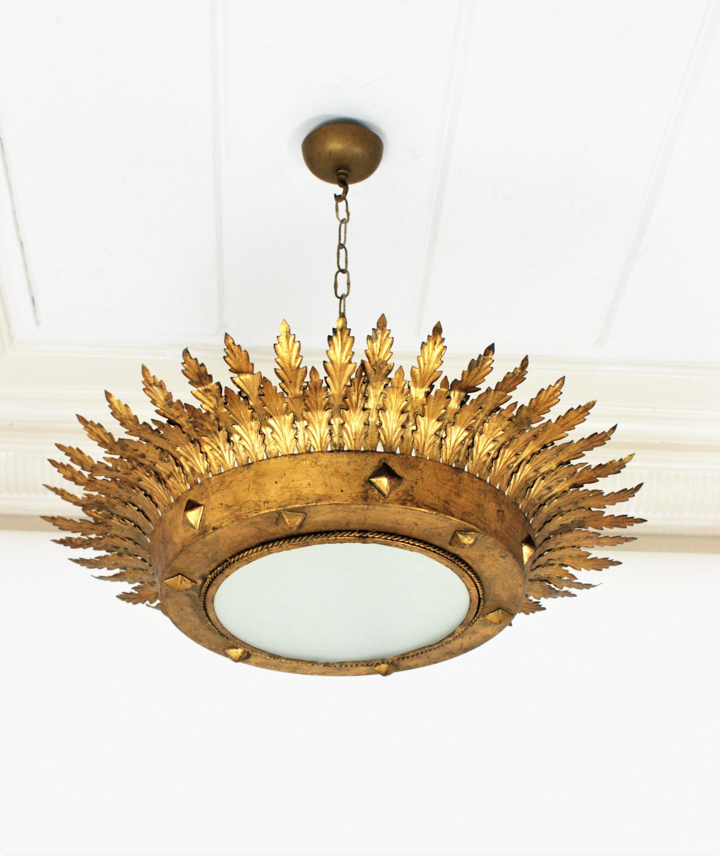 Dramatic large sunburst crown ceiling light fixture or chandelier / pendant with leafed frame and frosted glass diffuser. Spain, 1940s
This ceiling light was handcrafted in Spain and its design combines neoclassical and Hollywood Regency accents.