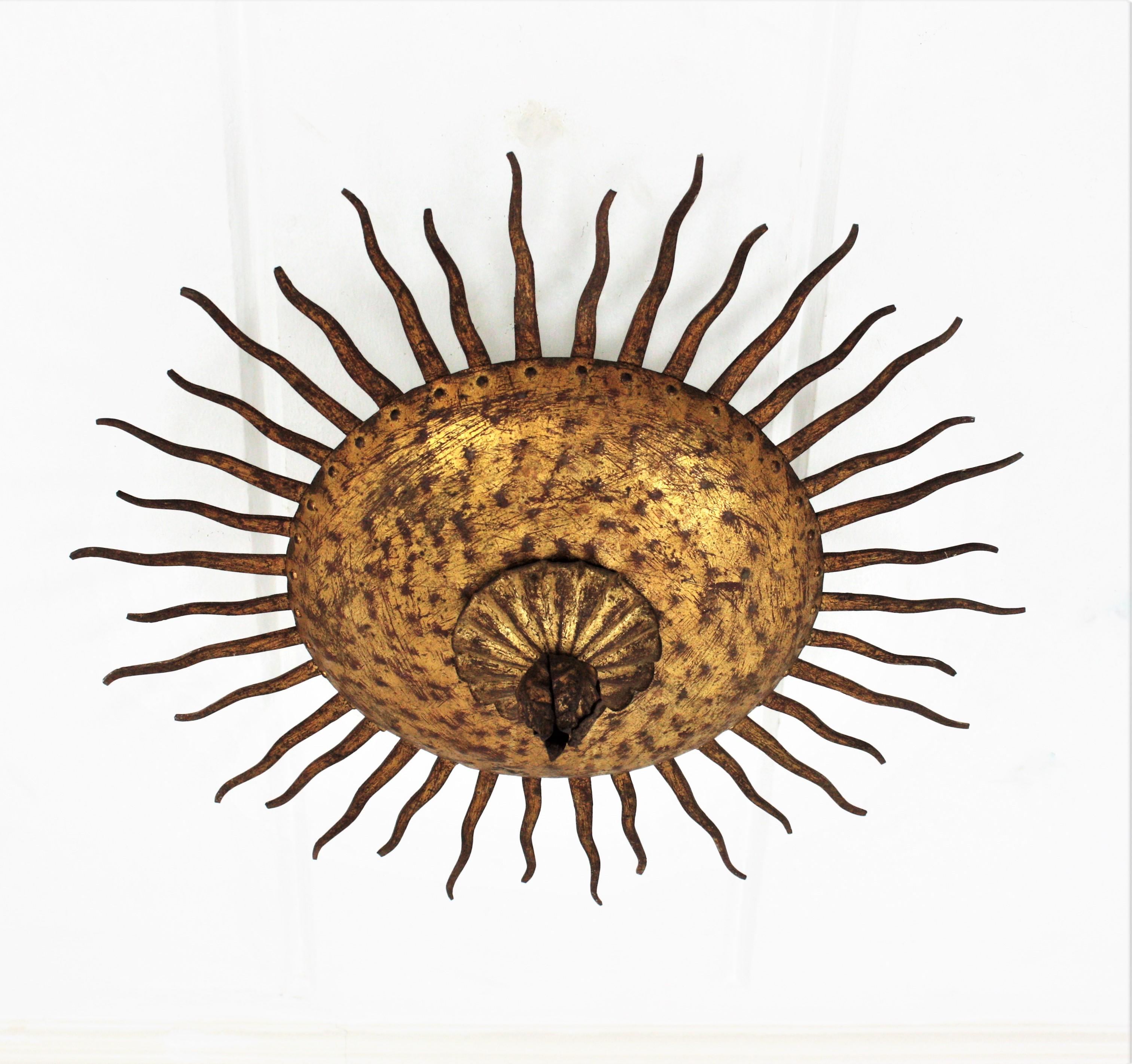 Wrought gilt hand forged iron sunburst flush mount / ceiling light fixture. France, 1950s.
This hand-hammered iron flushmount has alternating rays in two sizes surrounding the central sphere and a flower decoration at the central part. 
This