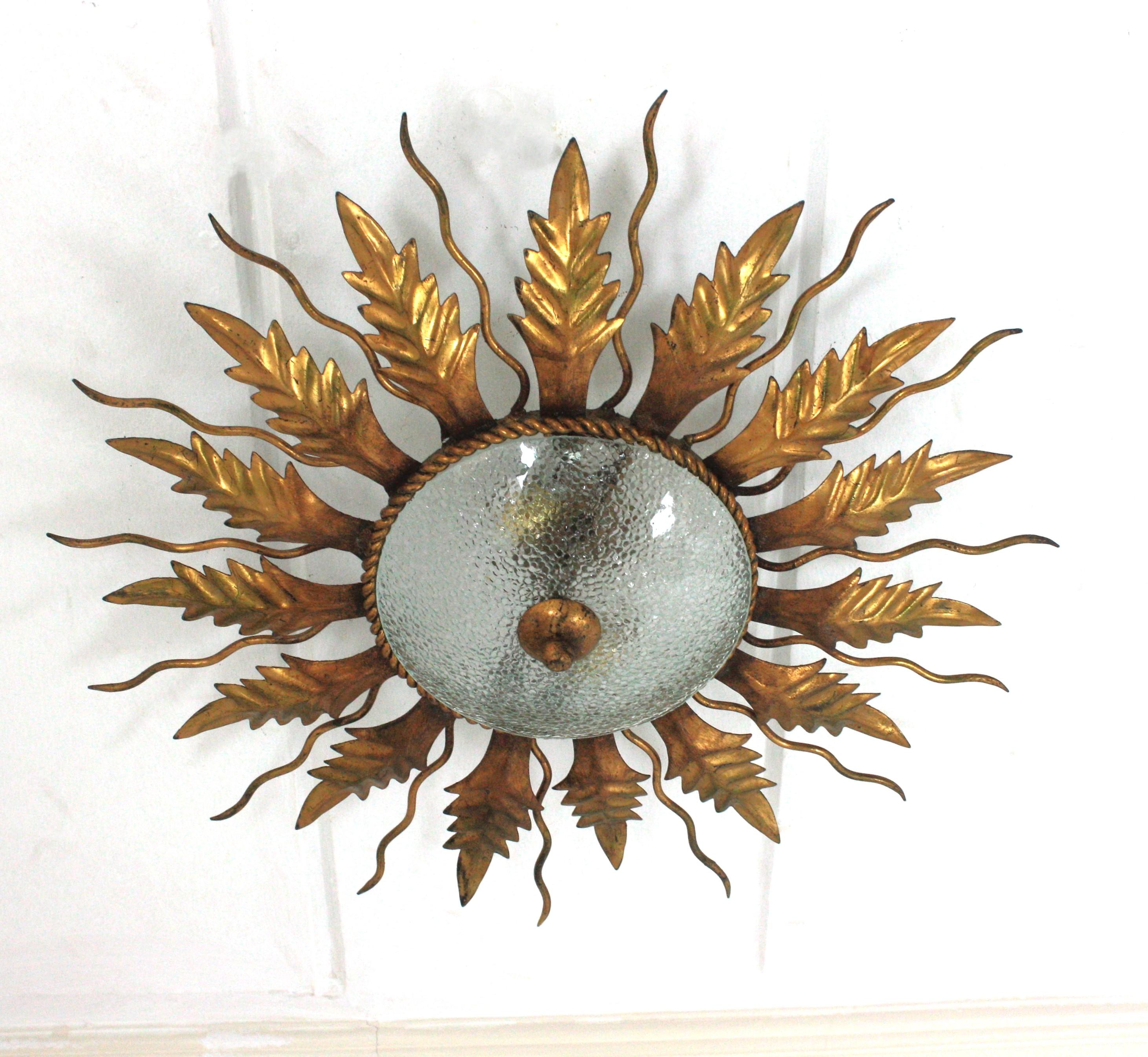 Gold leaf Gilt sunburst light fixture in wrought iron and cracked glass. Spain, 1950s.
This eye-catching ceiling flush mount is made in hand-forged iron and finished in gold leaf gilding.
It has curly iron rays alternating with iron leaves