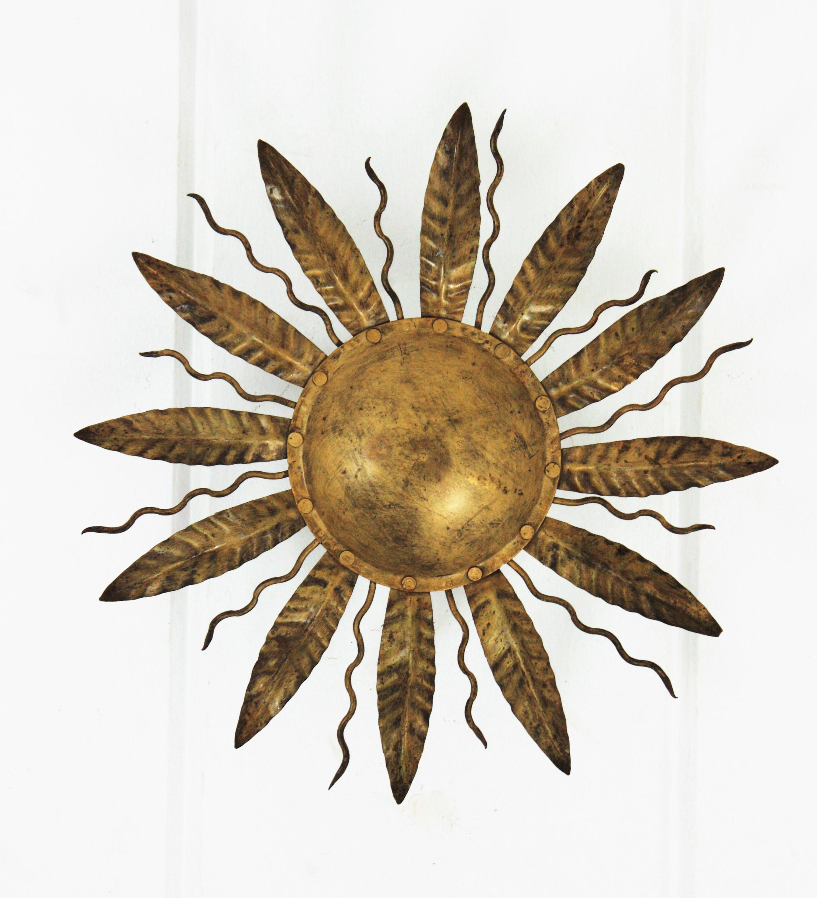 Lovely gilt metal leafed sunburst flushmount. Spain, 1950s-1960s.
This ceiling flushmount features an iron structure with a central round panel surrounded by alternating leaves and curly rays.
It has a nice aged patina.
Two E14 bulbs at the back