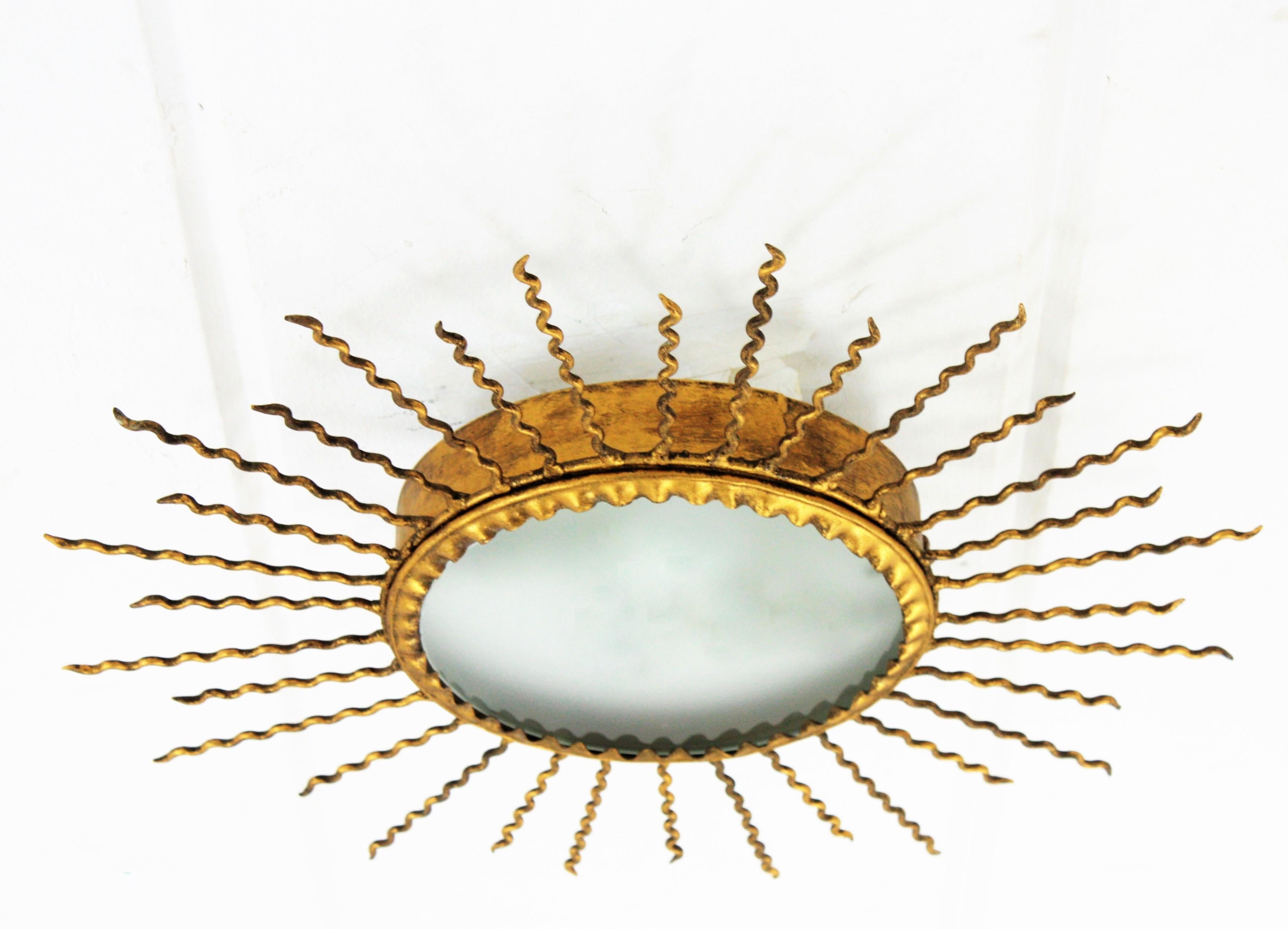 Spanish sunburst flush mount or mirror in gilt iron with curly rays, 1950s.
Wrought iron sunburst light fixture mirror with curly rays and frosted glass diffuser. It can be used as ceiling light fixture, as wall sconce and as sunburst mirror or