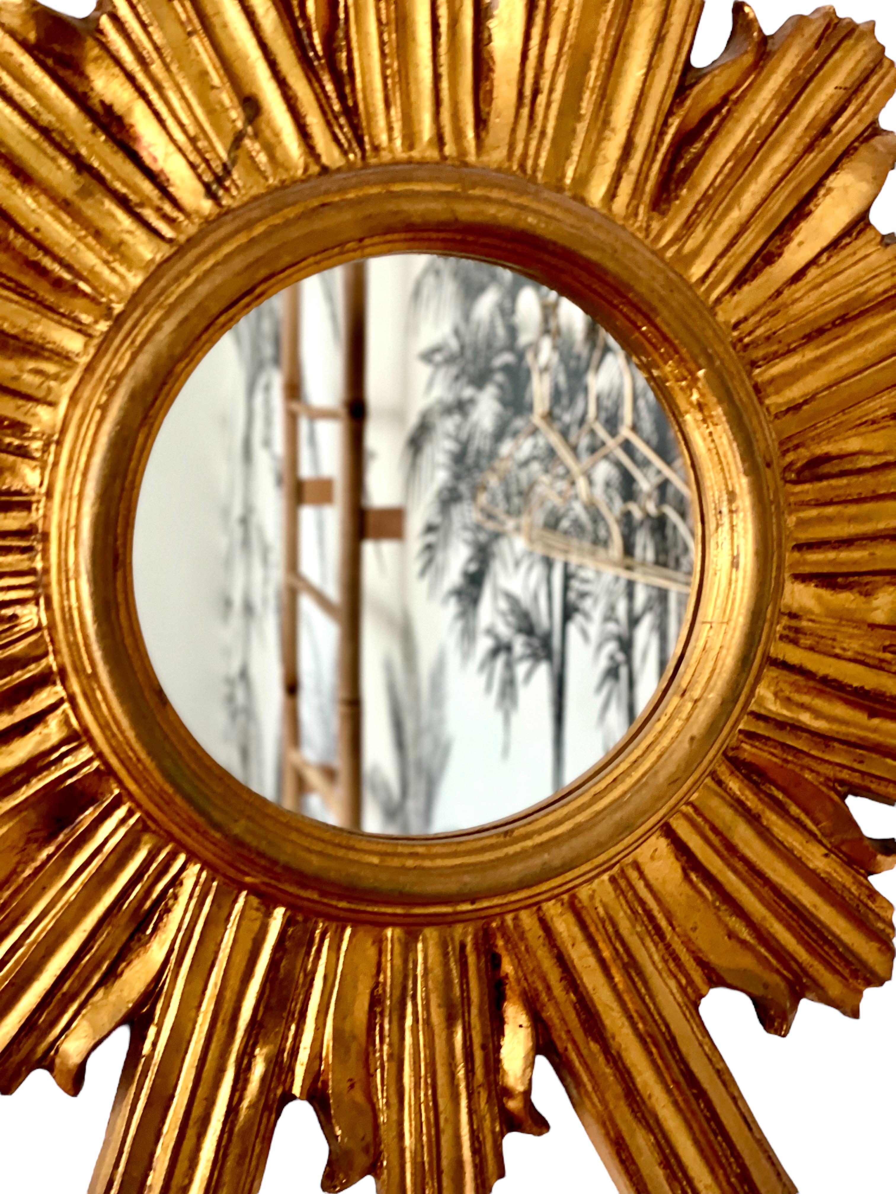 A gleaming Florentine-style antique sunburst mirror in carved and gilded wood, beautifully handcrafted in the mid-20th century. The small, circular mirror plate is enclosed by an array of carved golden rods, some straight, and some undulating, which