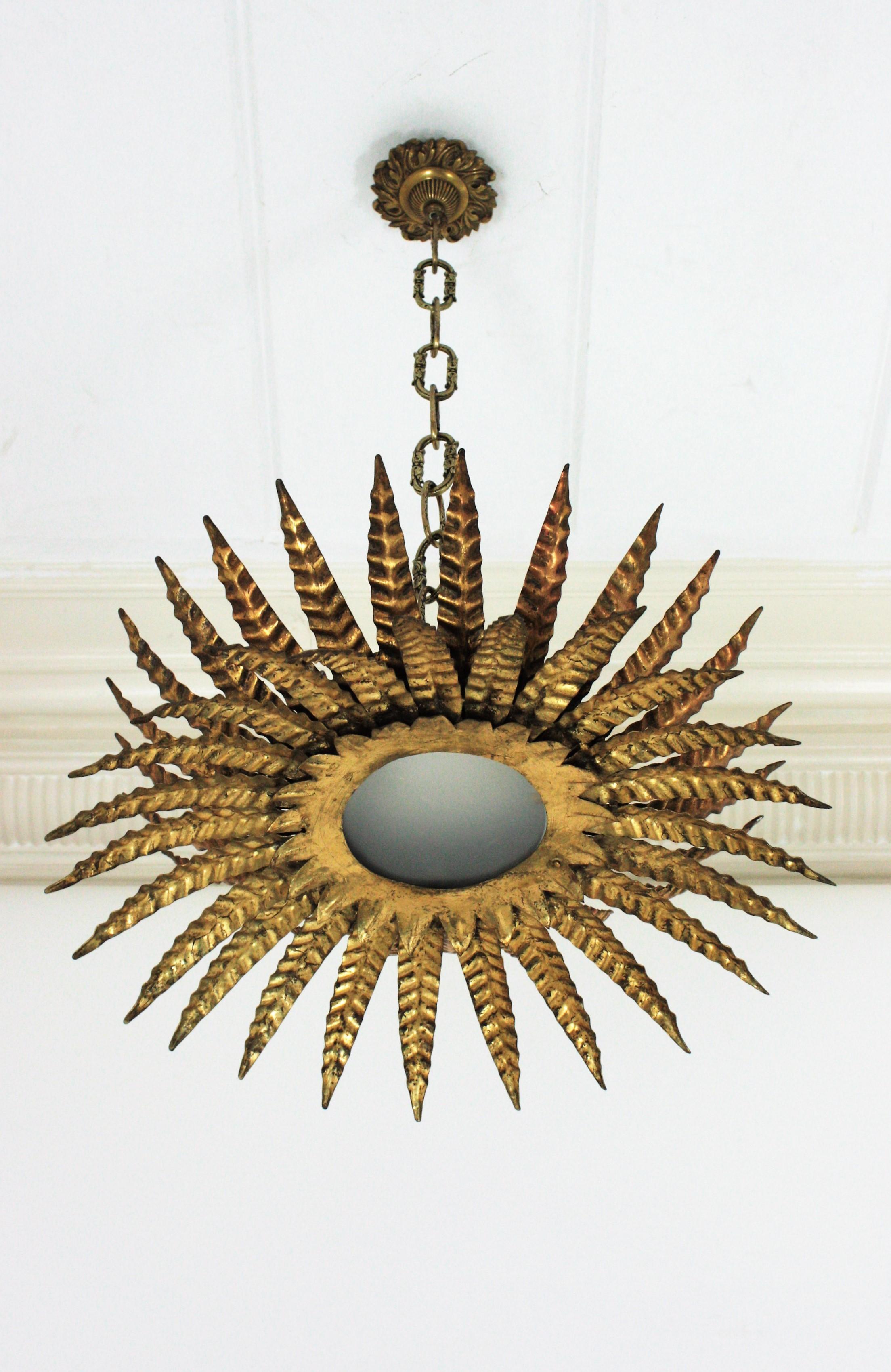 Gorgeous Brutalist gilt iron and glass sunburst flush mount ceiling lamp or chandelier. France, 1950s.
This sunburst light fixture features a triple layer of leaves / rays surrounding a central frosted glass panel. Each layer has rays in different