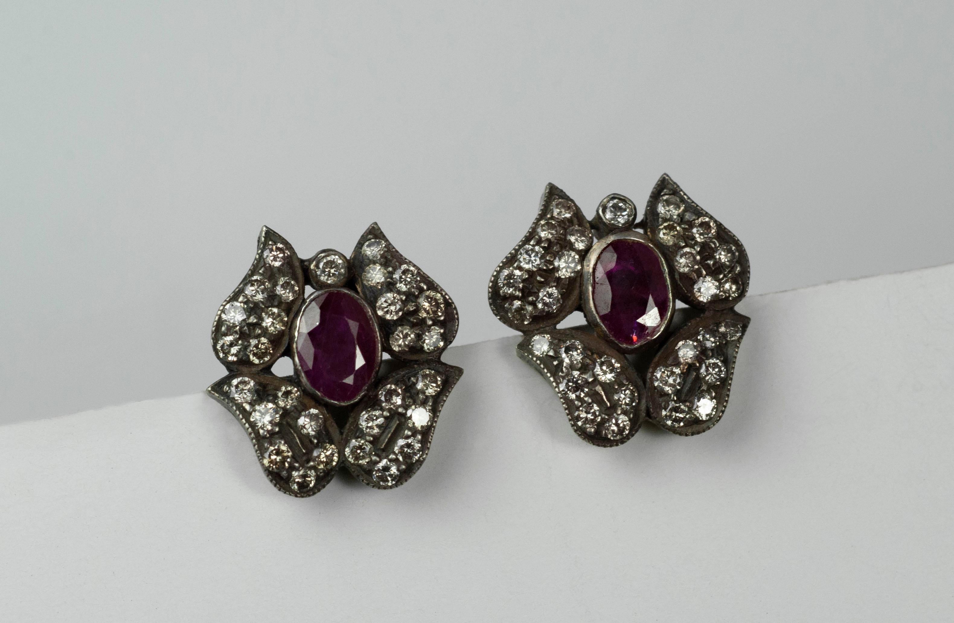 Unique stud earrings in oxidized sterling silver featuring a faceted oval ruby center and pave diamonds, with 14K gold posts.