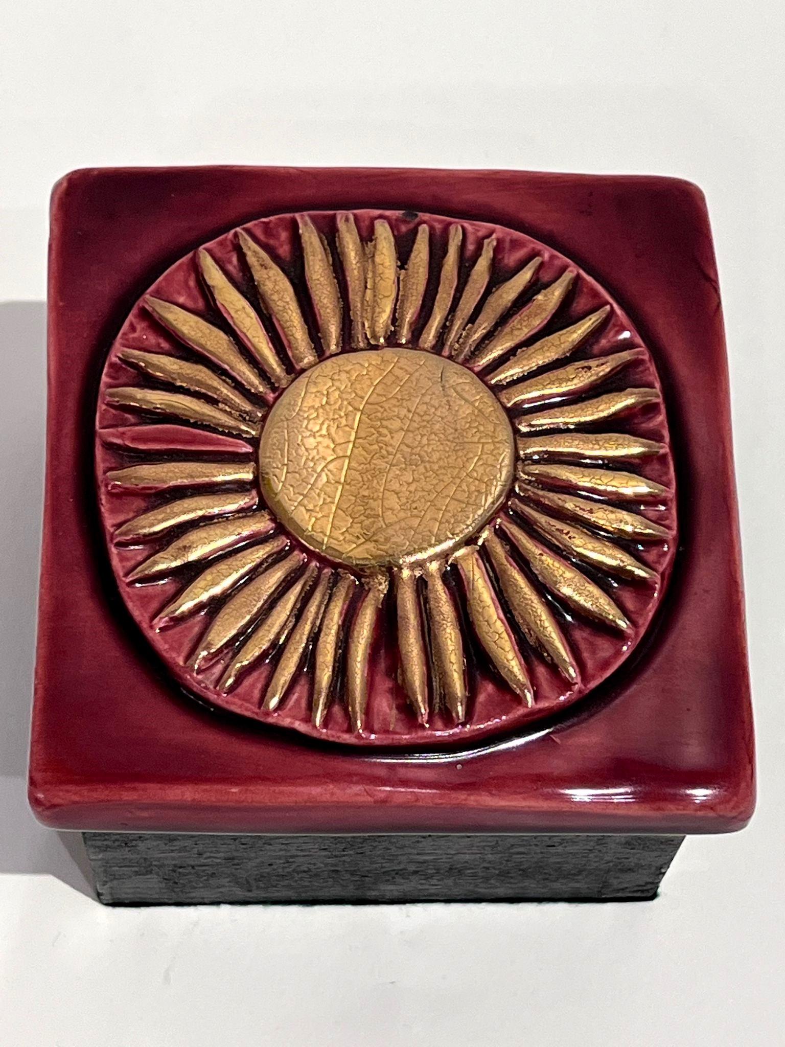Leichter Sprung in der Emaille an einer Ecke

Das Holzkästchen wird von einem Deckel aus geprägtem Steingut gekrönt, der mit burgunderroter Emaille und einer goldenen Sonnenblume verziert ist und eine kostbare warme und leuchtende Harmonie bietet.